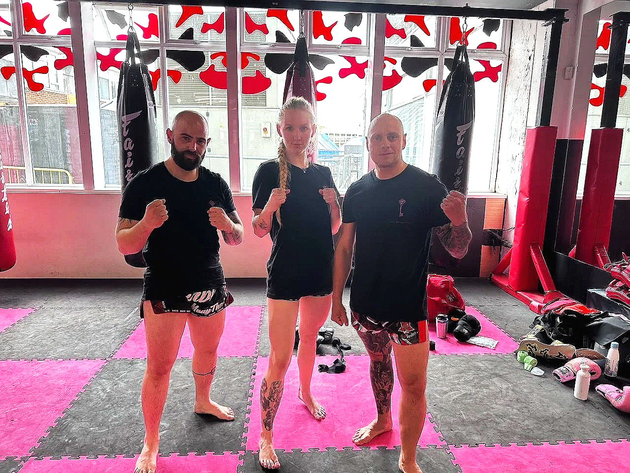 Three people in martial arts training gear standing inside a gym, posing with fists raised. Two men and one woman are barefoot, surrounded by punching bags, gloves. The gym has red and black flooring with pink and red wall decorations.