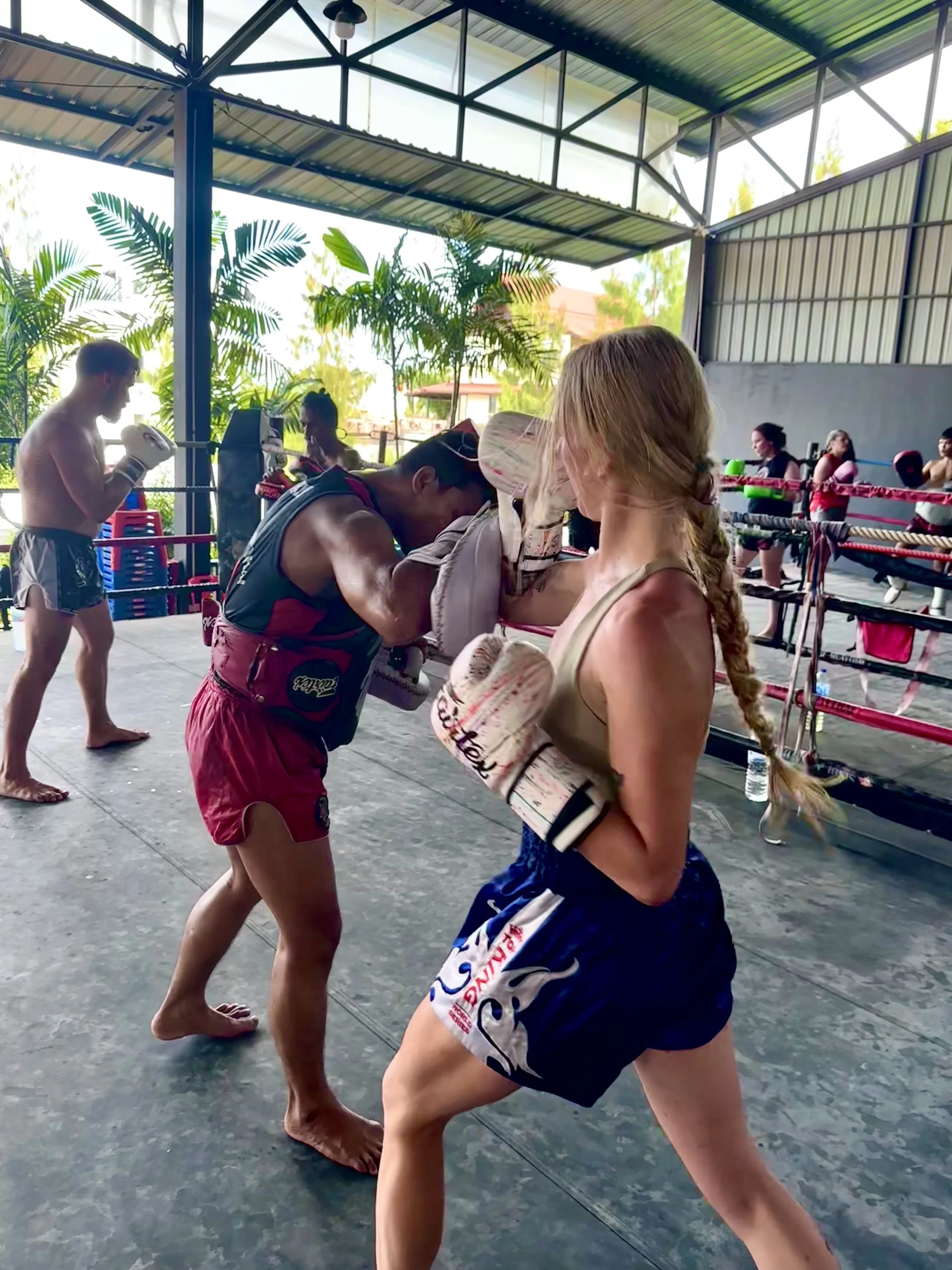 A woman practicing boxing in a gym with trainers, while other martial artists train nearby.