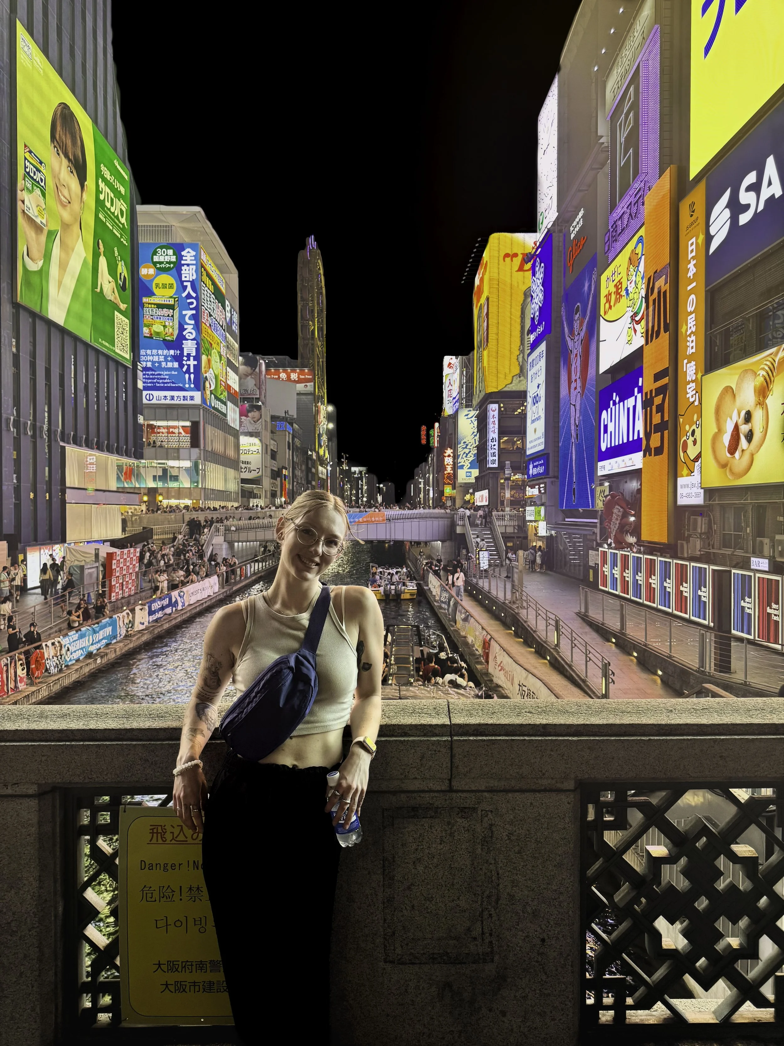 A woman standing in front of a canal at night in an urban area with bright billboards and neon lights, smiling at the camera.