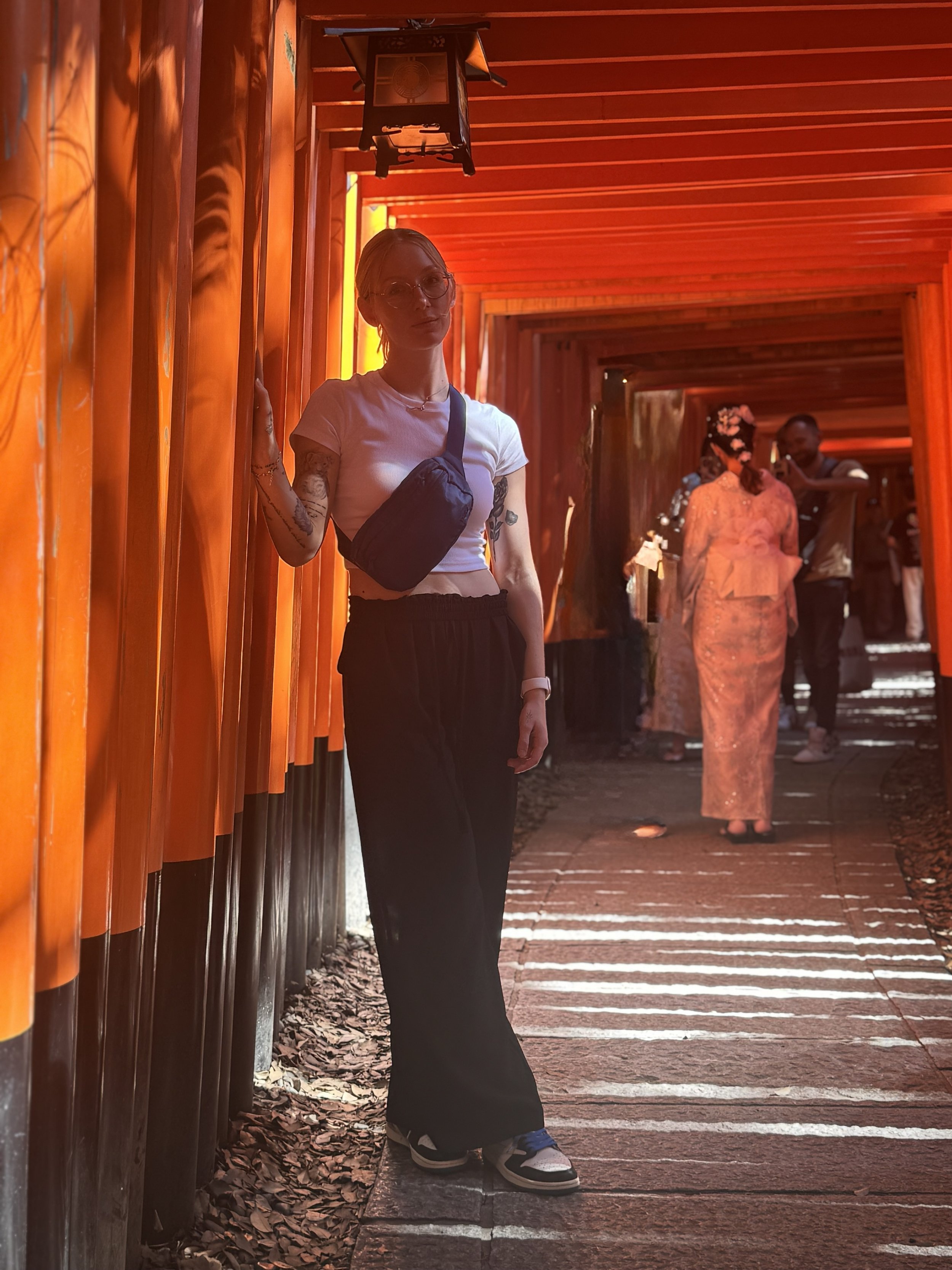 Person standing near orange-colored traditional Japanese torii gates with other people in traditional attire in the background.