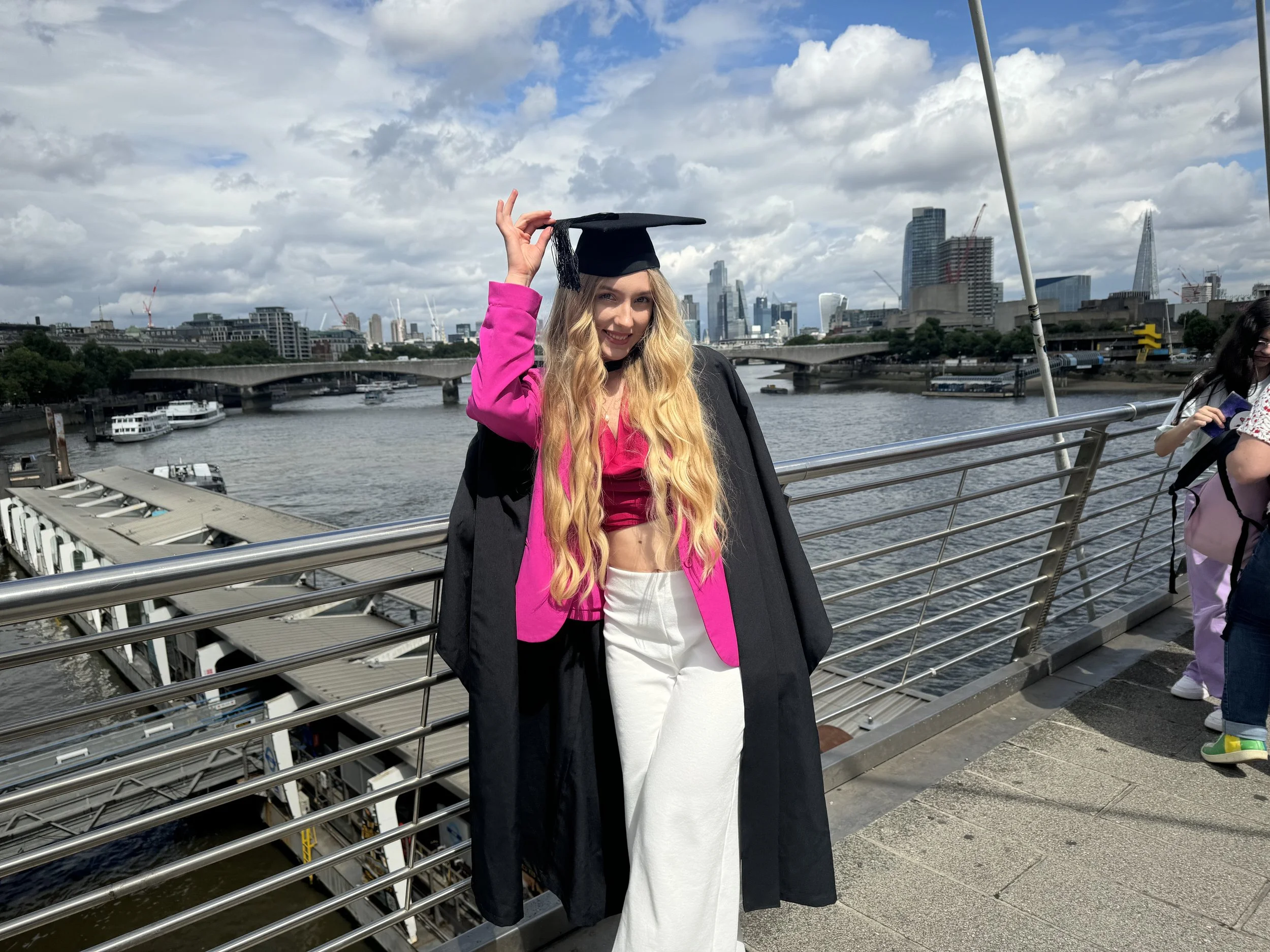 A young woman in graduation attire, with a pink blazer and white trousers, stands on a bridge over the river Thames in London. She is holding her graduation cap and smiling, with a city skyline in the background.