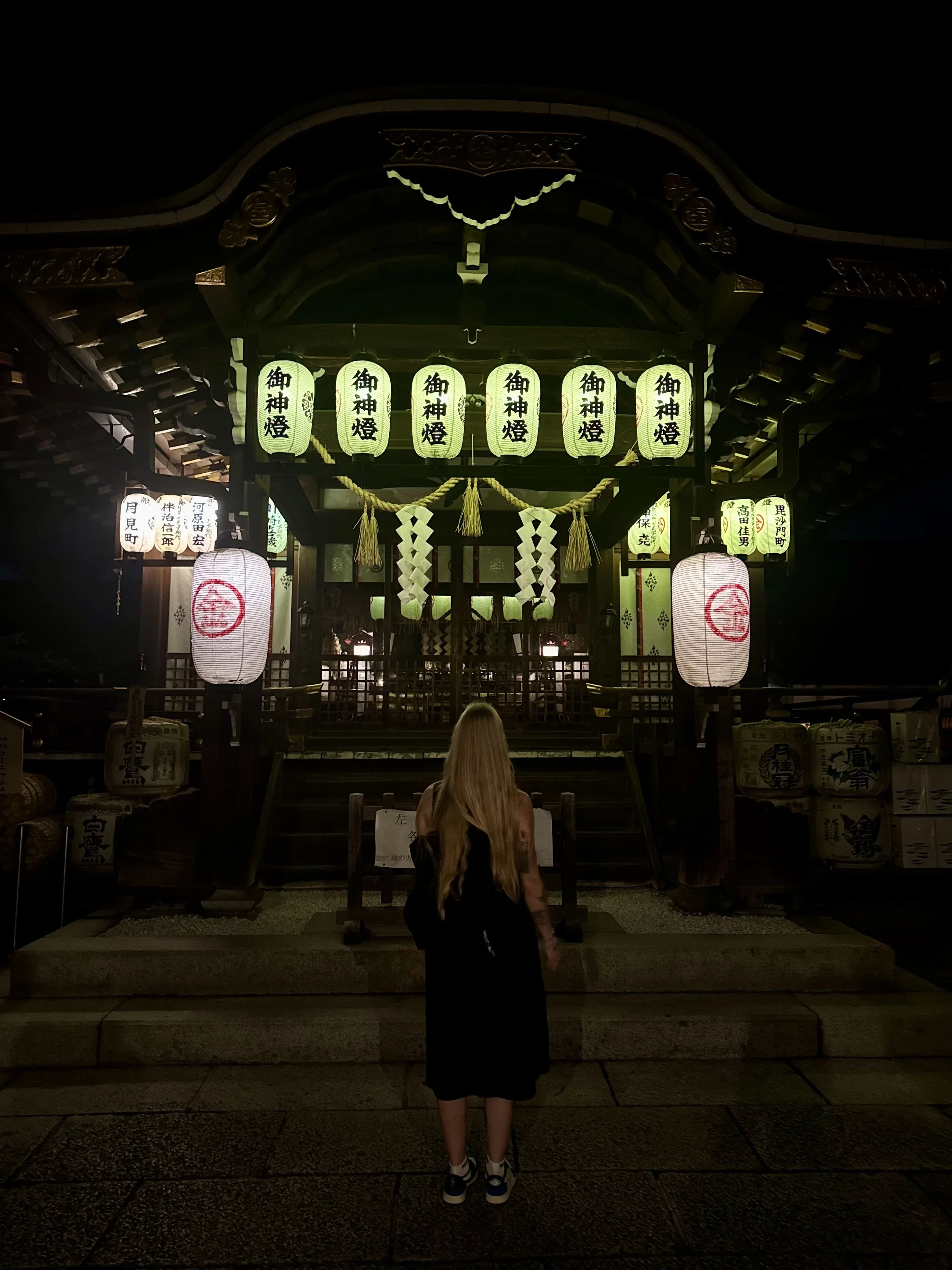 A woman with long blonde hair and a black dress standing in front of a Japanese shrine at night, illuminated by lanterns.