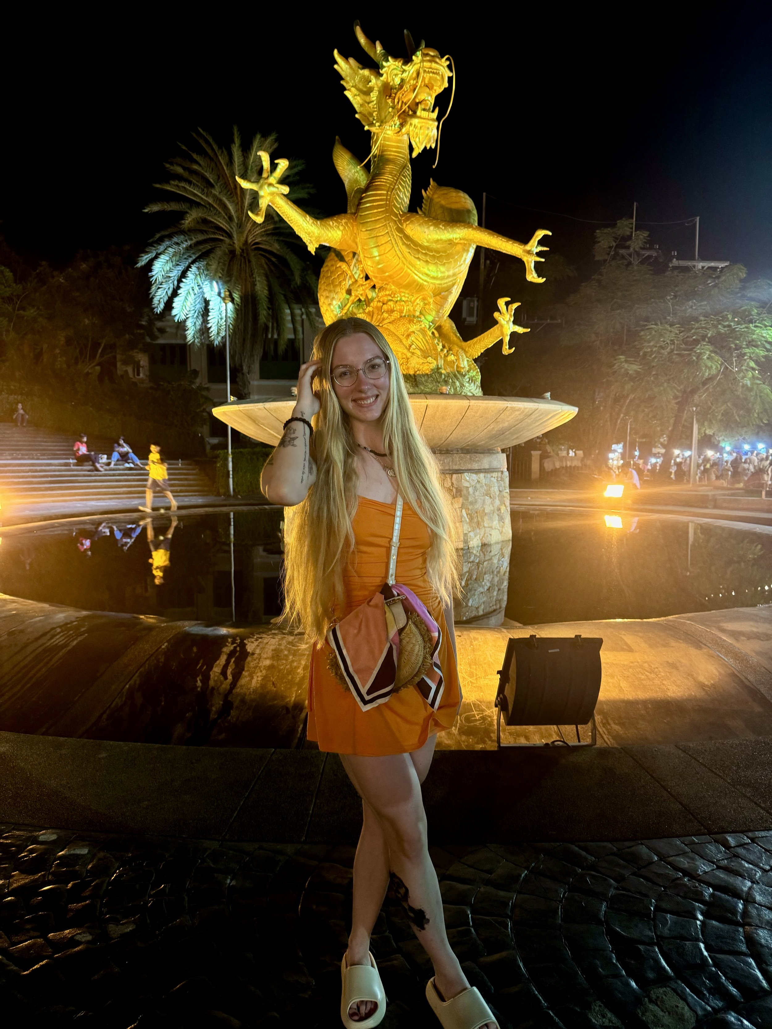 A smiling young woman with long blonde hair and glasses standing in front of a large golden dragon fountain at night. She is wearing an orange dress, beige slide sandals, and has a scarf tied around her bag.