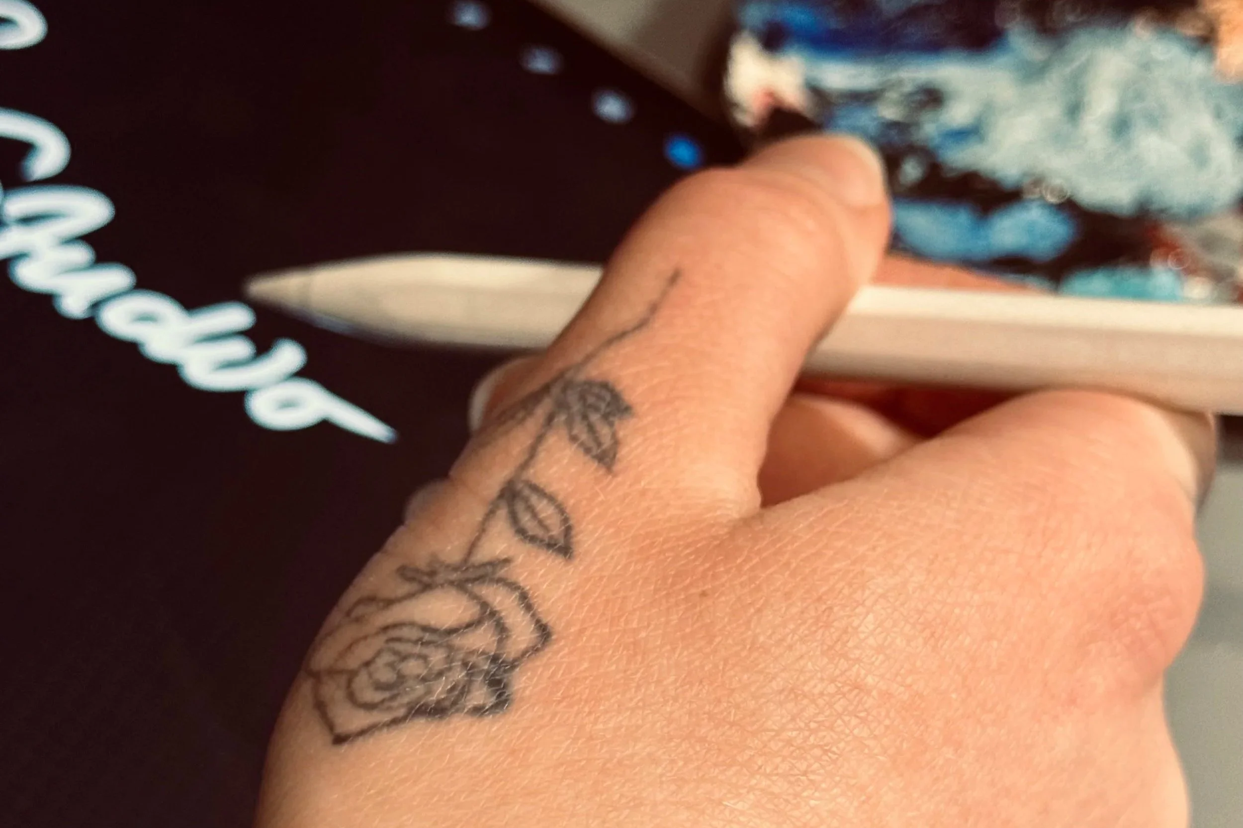 Close-up of a person's hand holding a digital pen, with a tattoo of a rose and leaves on the finger. In the background, part of the iPad is seen with the logo of the artist.