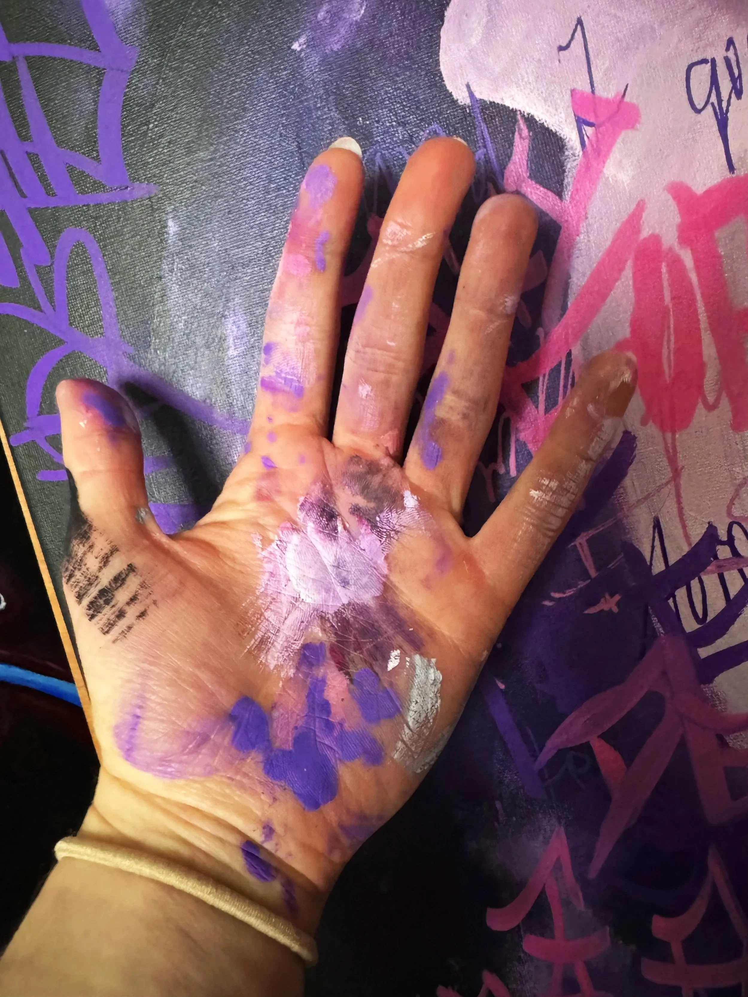 A hand smudged with purple, pink, and white paint rests against a painted surface with purple and pink graffiti-style artwork.