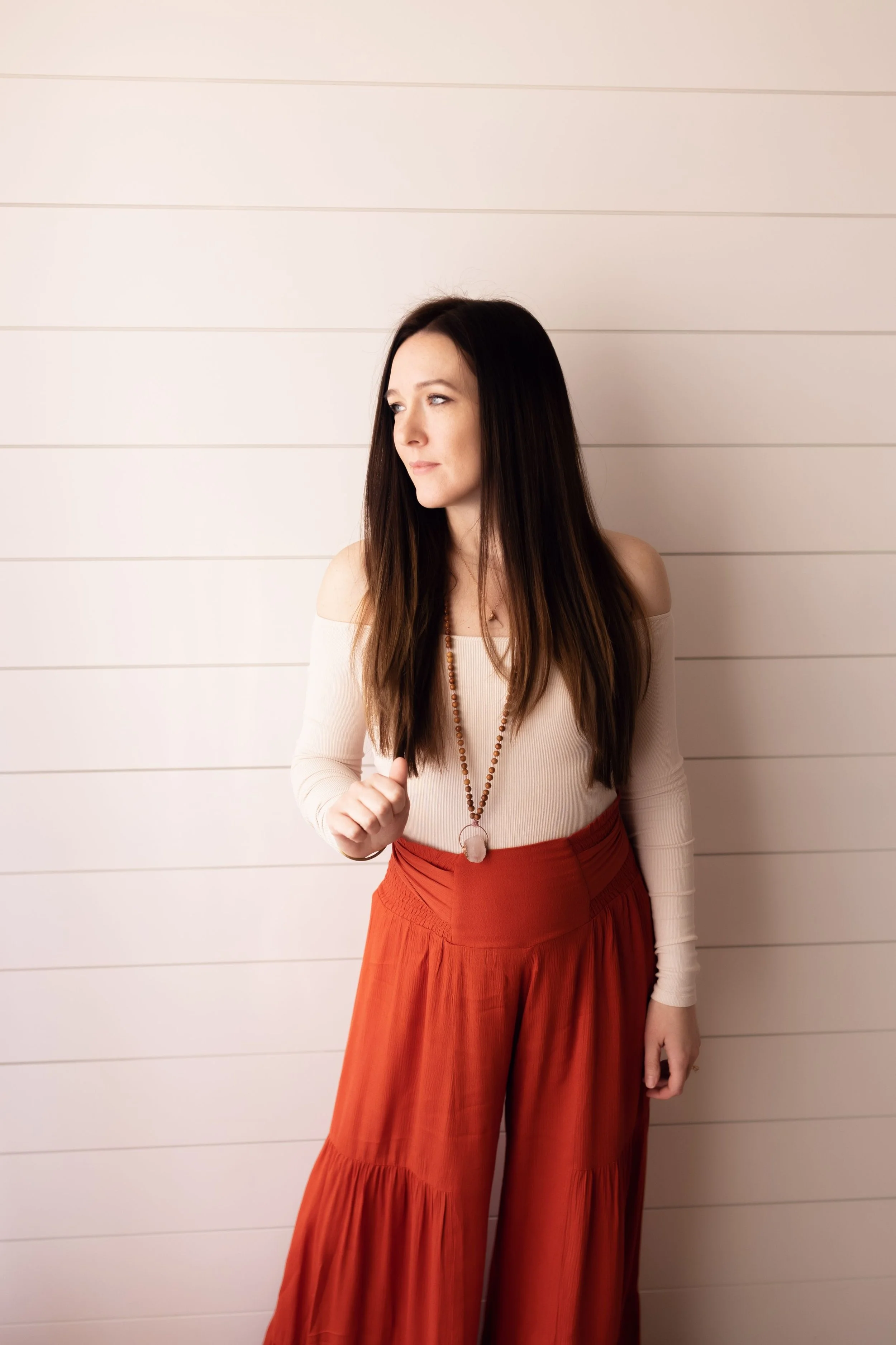 Crystal Energy Healer and Reiki expert, Jessica Blaess, is wearing a cream off-the-shoulder top, a beaded necklace, and wide-leg rust-colored pants, standing against a white paneled wall.