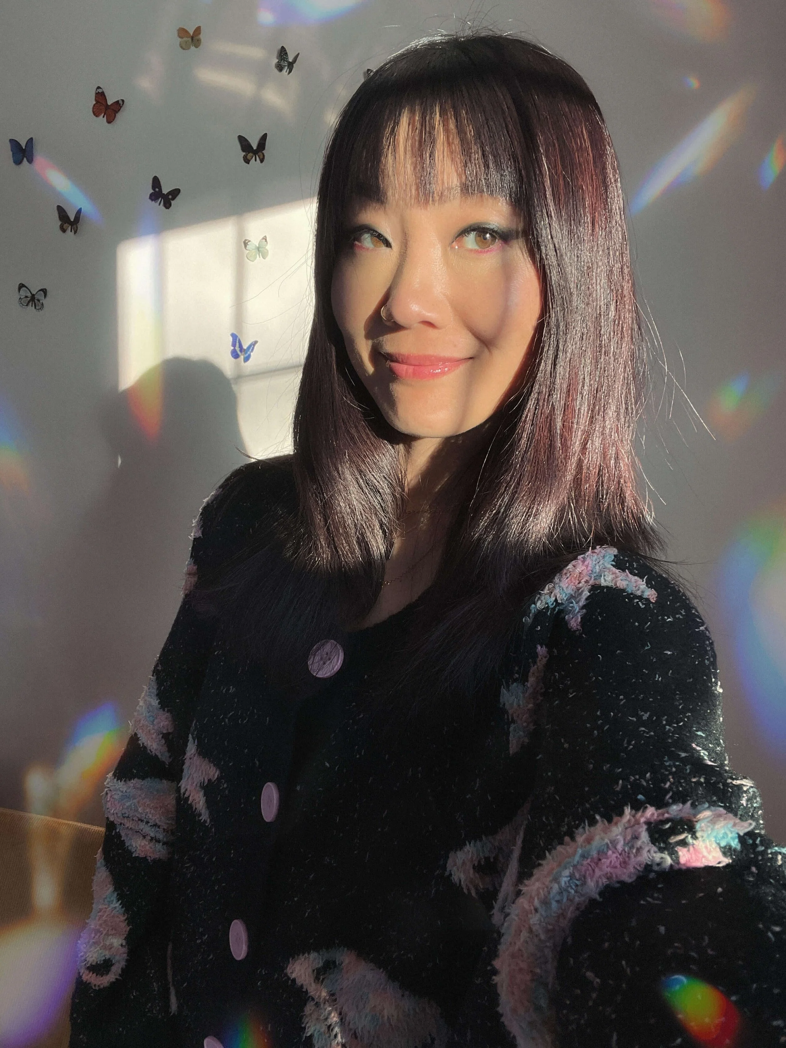 Psychic certification program student, Corey Mee, smiles while wearing a black top with stars on it, the wall behind her adorned with butterfly stickers and rainbow prisms on the walls from the window light in front of her.