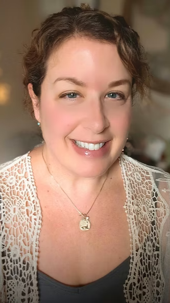 Professional Numerologist, Kimberly Beauchemin, sits with short, curly brown hair smiling at the camera, wearing a lace top and a necklace with a pendant.