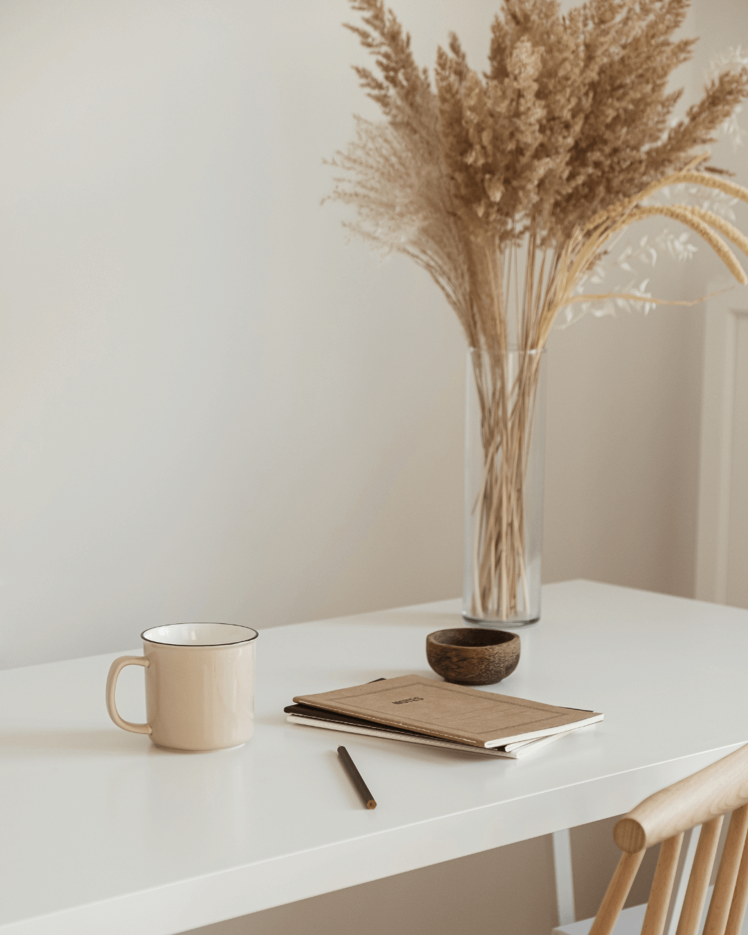 A minimalist white table with a beige coffee mug, a closed notebook, a pen, and a small wooden bowl, with a tall glass vase of dried pampas grass in the background.