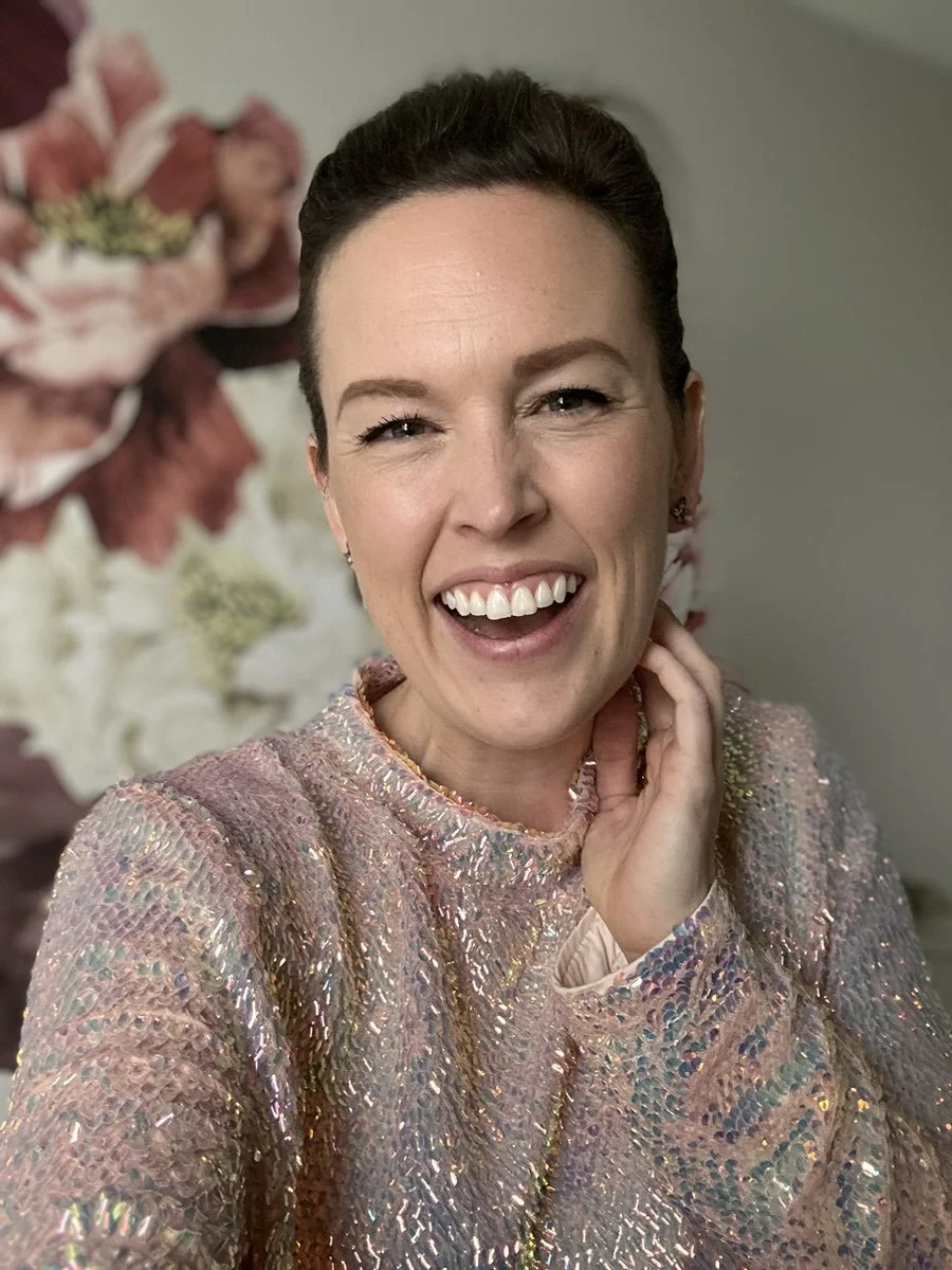 Psychic Development student, Sarah-Beth B. smiles as she touches her face with her left hand, wearing a sparkly, pinkish, sequin-like top, standing in front of a floral background with large, out-of-focus pink, white, and green flowers.