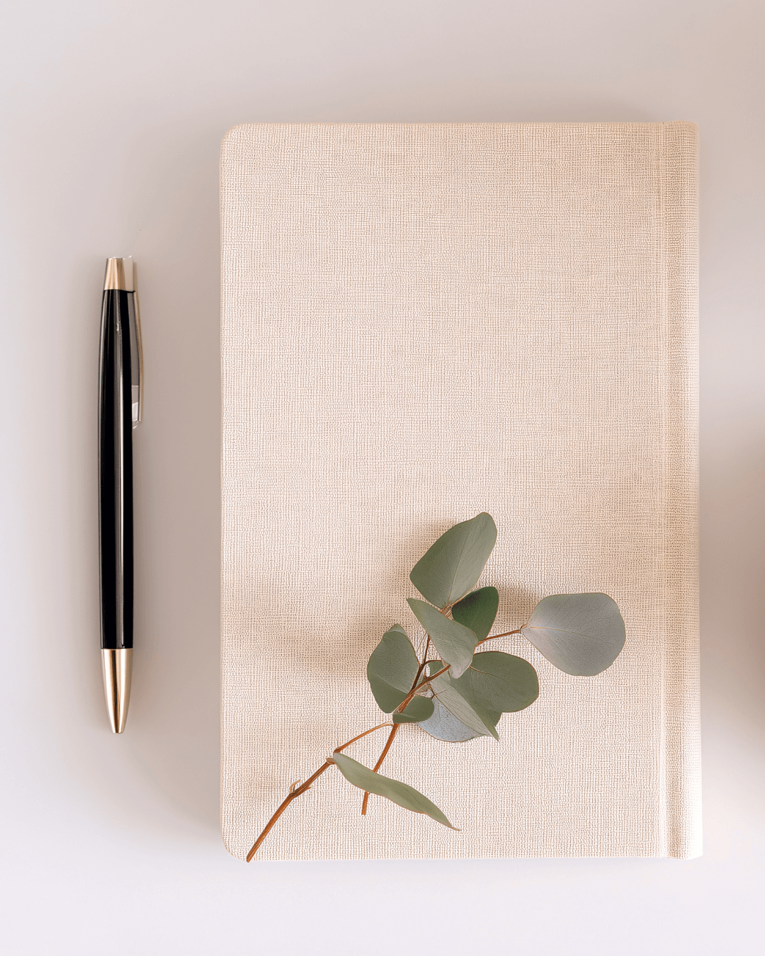 A beige fabric-covered notebook with a sprig of eucalyptus leaves on top, a black and gold pen on the left side, on a white surface.