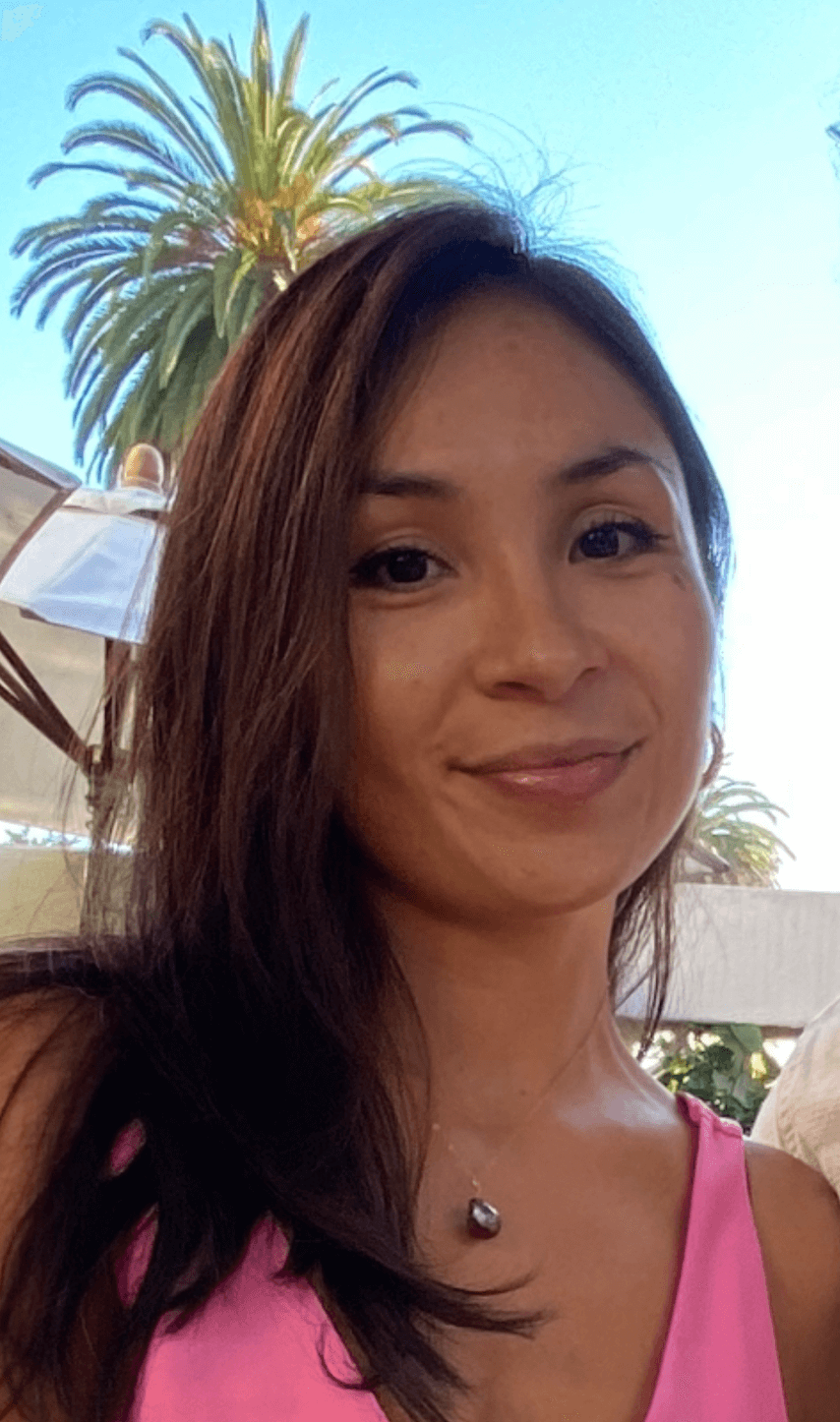 Psychic Development student, Molly L., is wearing a pink top and a necklace with a dark pendant, smiling outside with a palm tree and clear blue sky in the background.