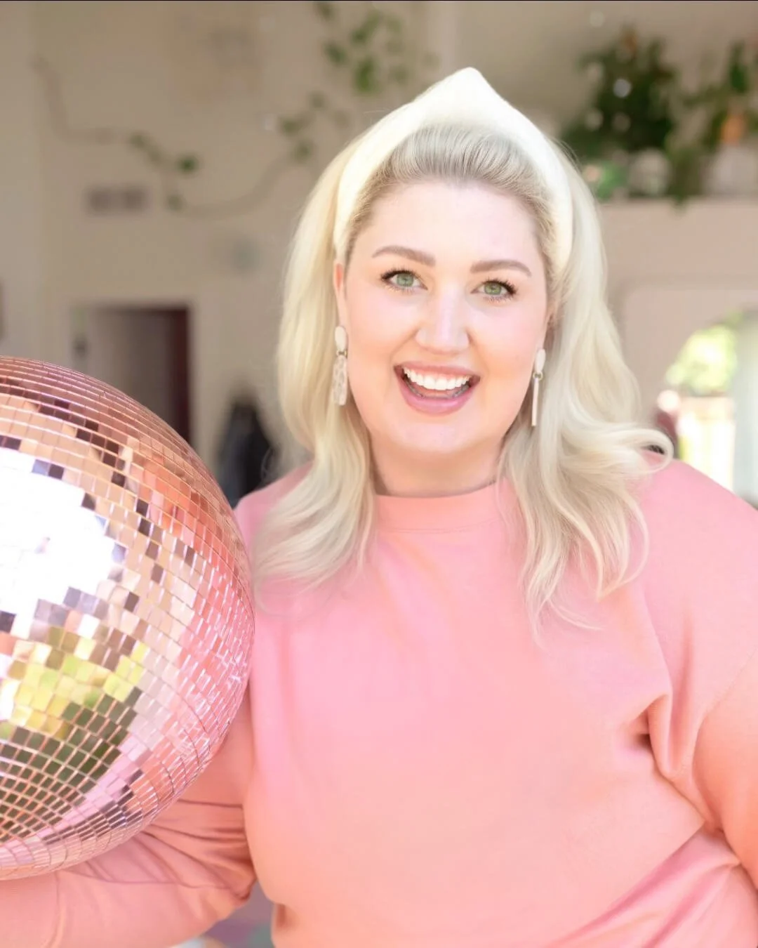 Psychic certification program student Bunny W smiles next to a large pink disco ball while wearing a pink sweater, cream headband, and dangling earrings.