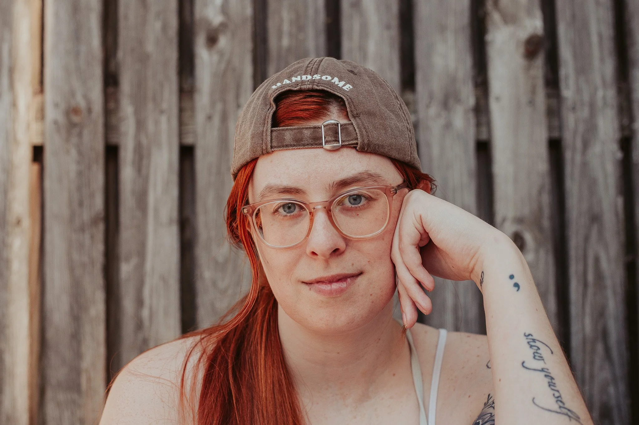 Psychic certification program student, Kelsey G, is wearing a backwards hat and red-rimmed glasses as sits smiling and rests her hand on her cheek.