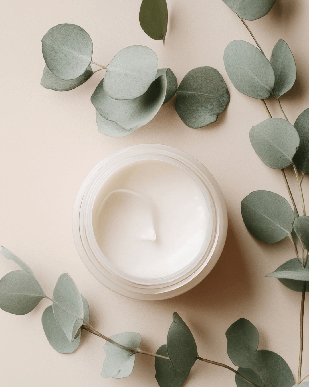 A white jar of cream surrounded by eucalyptus leaves on a beige background.