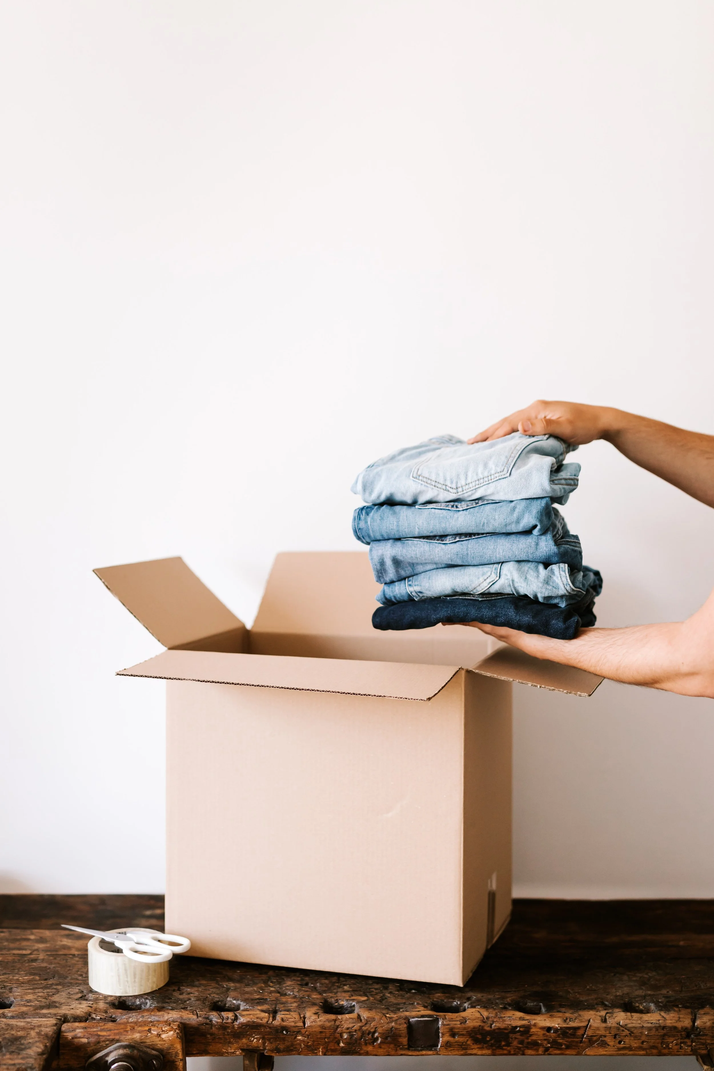 Person packing a cardboard box with folded jeans, on a wooden surface with scissors and tape nearby.
