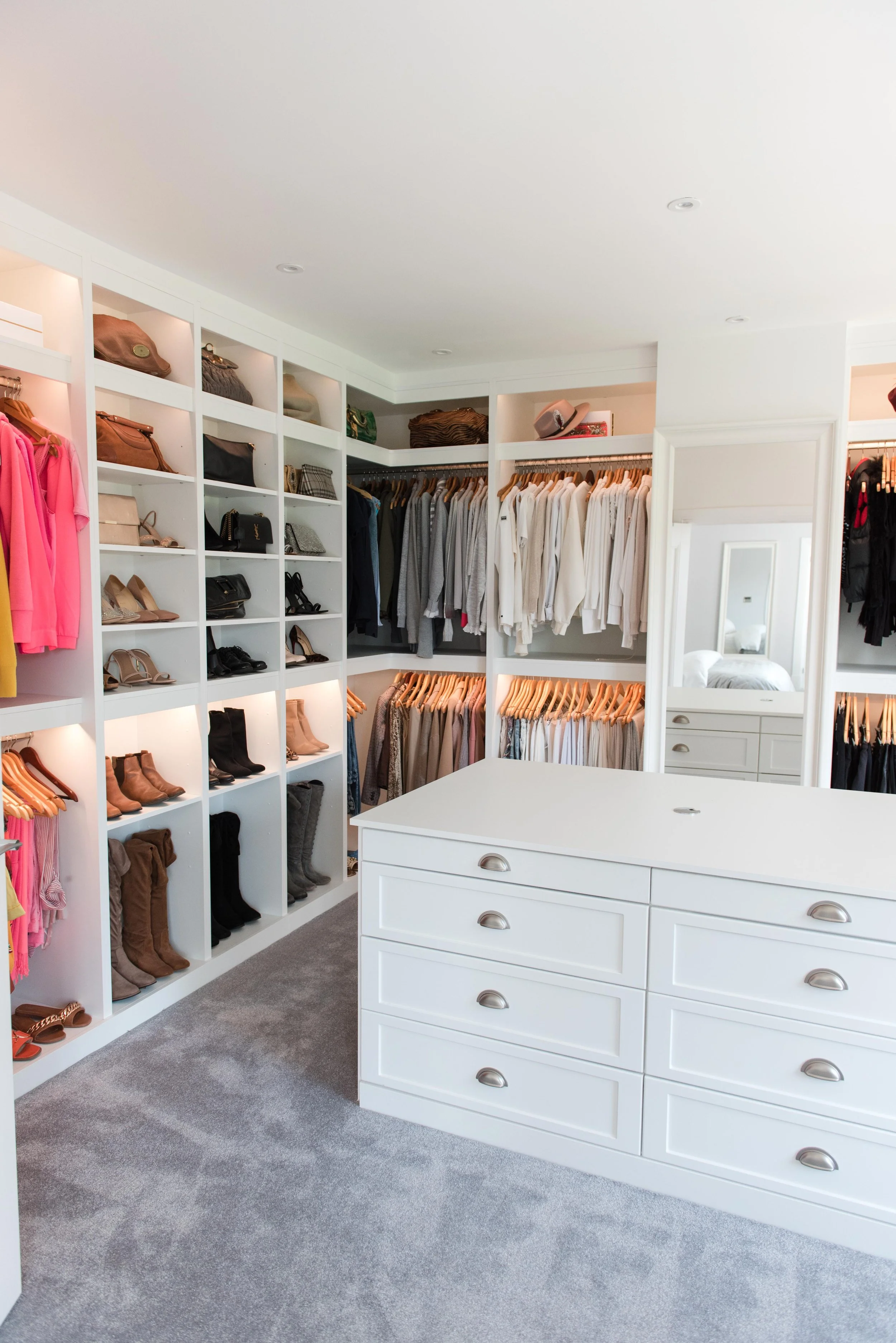 A walk-in closet with white shelving and drawers, filled with handbags, shoes, clothes, and hats, and a large mirror.