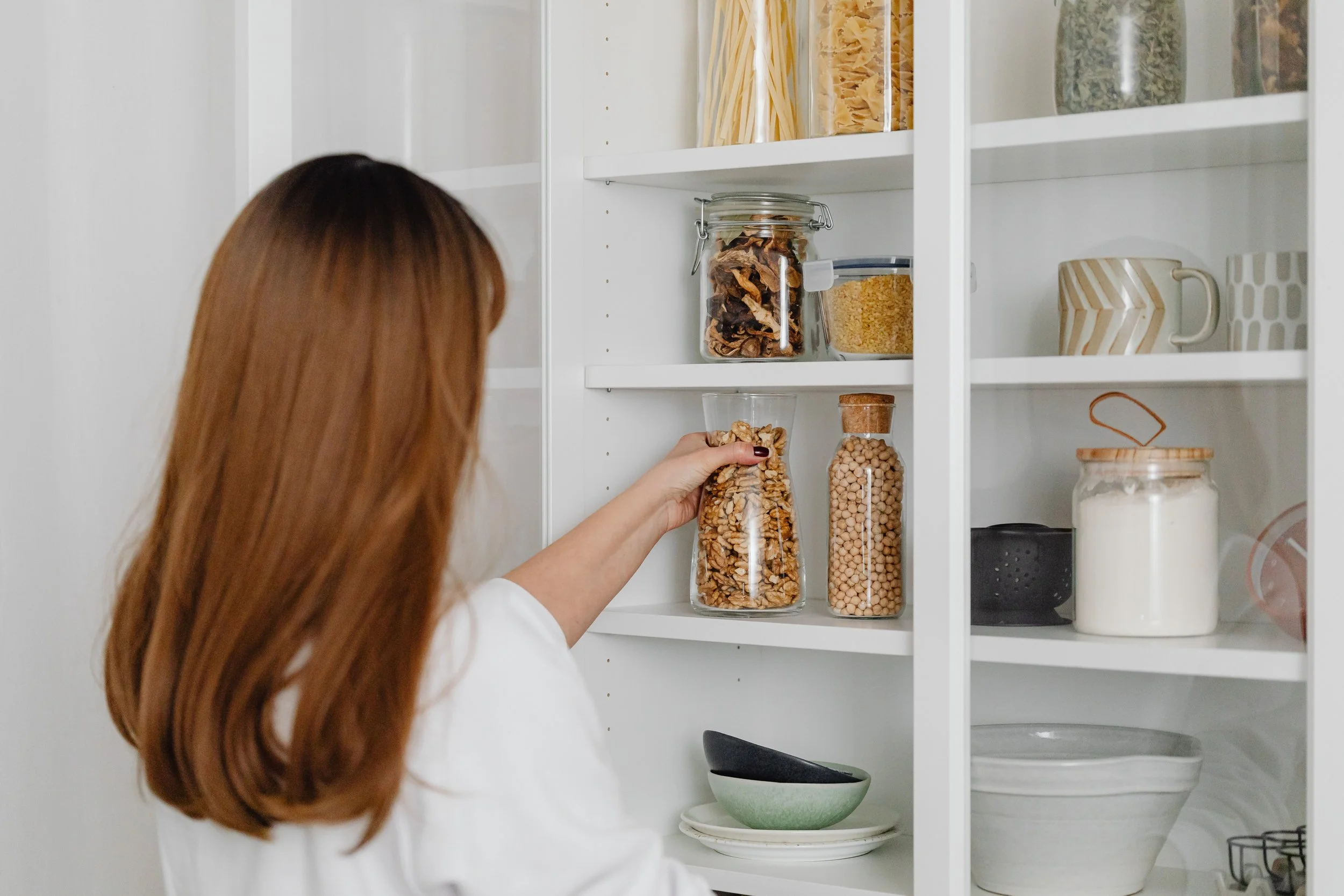 A woman with long brown hair reaching into a white pantry to grab a glass jar filled with nuts. The pantry shelves contain various labeled jars with dry foods such as pasta, dried mushrooms, and chickpeas, along with bowls and a jar of flour.