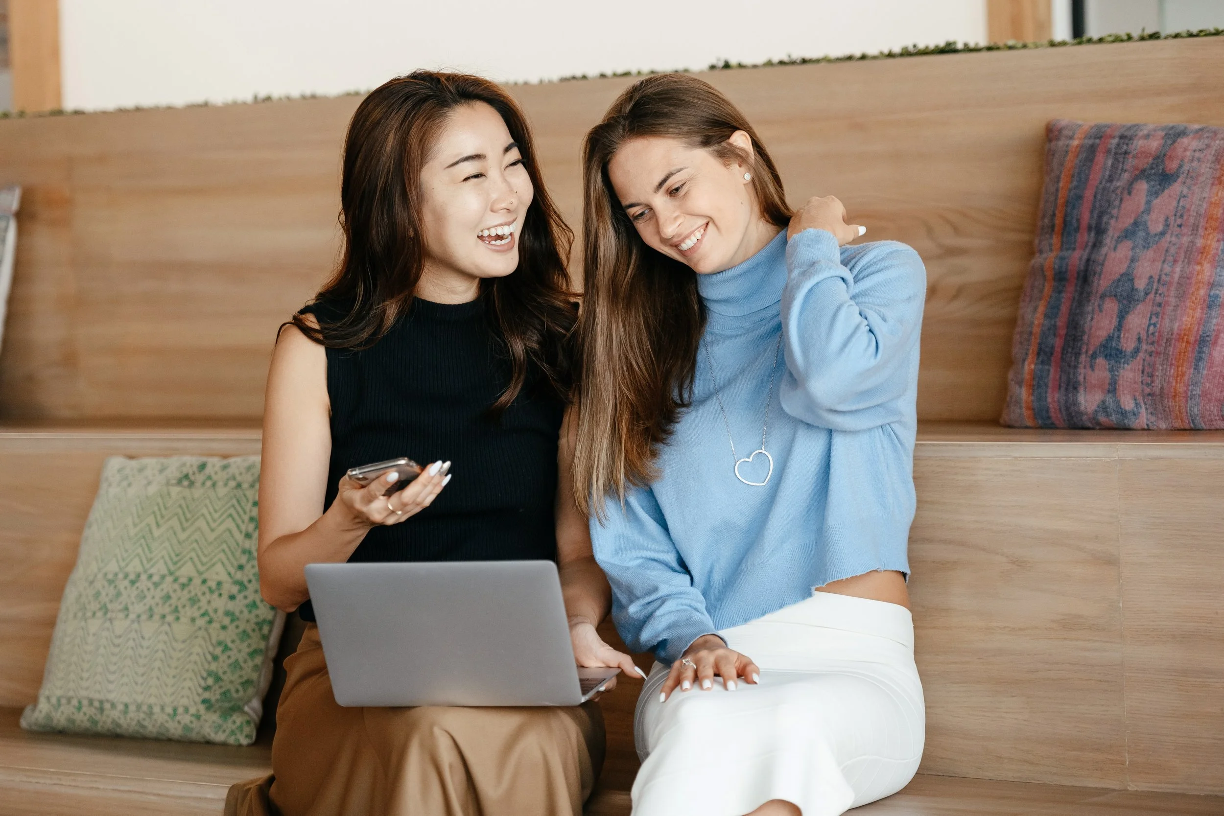 Two women sitting on a wooden bench, smiling and laughing, one holding a smartphone and a laptop, in a cozy indoor setting with decorative pillows around them.