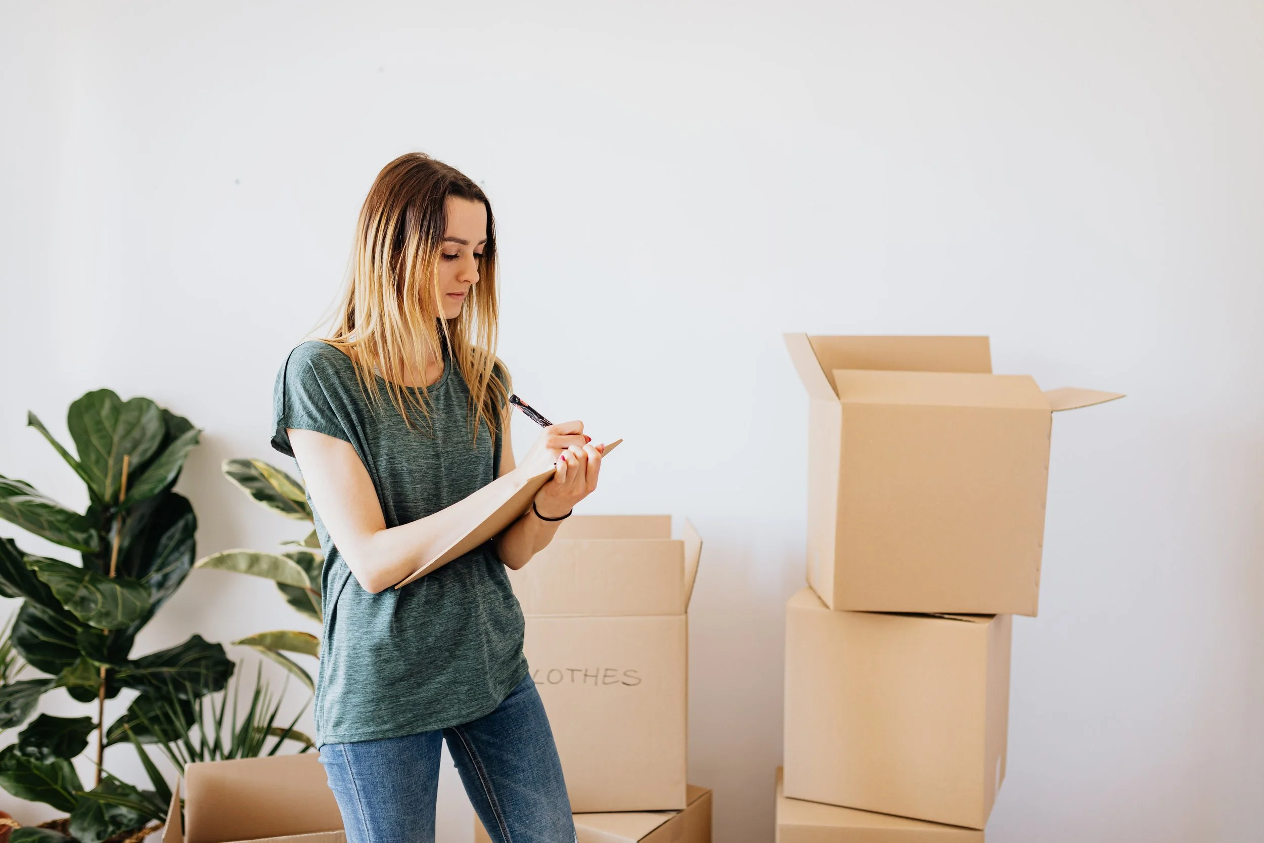 A young woman with long blonde hair wearing a green t-shirt and jeans, writing on a clipboard, surrounded by moving boxes and a large plant.