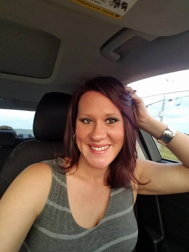 A woman with shoulder-length auburn hair, wearing a sleeveless gray striped top, smiling and sitting in a car.