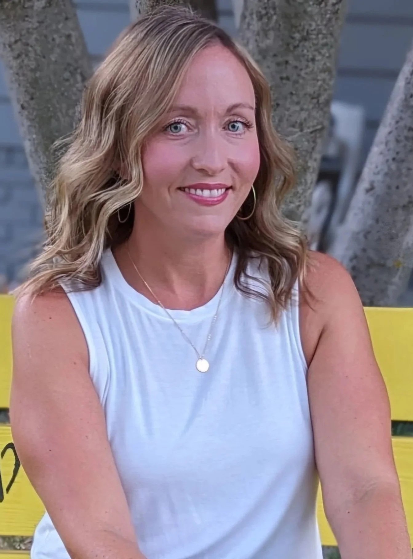 A woman with blonde wavy hair, wearing a white sleeveless top, sitting on a yellow bench outdoors, smiling, with trees and a body of water in the background.