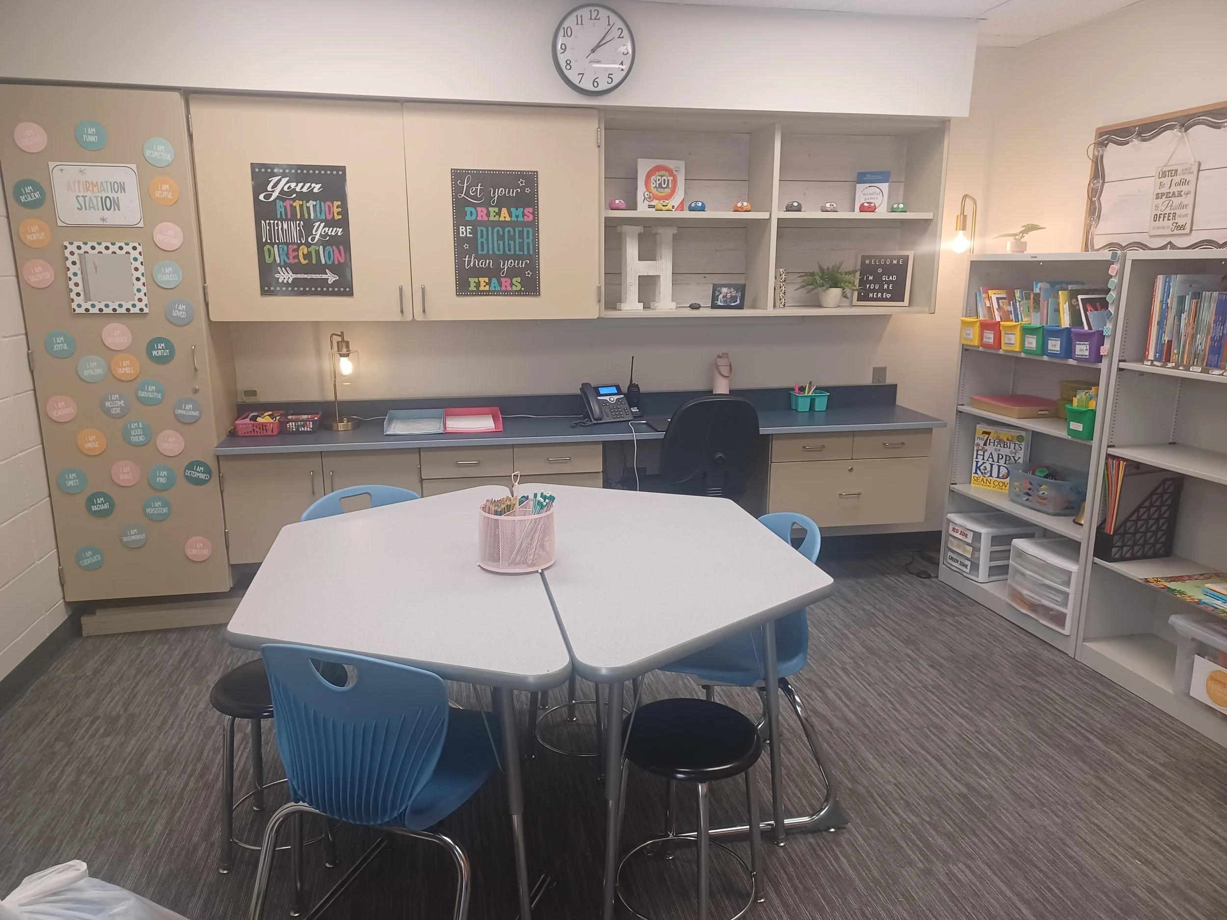 A classroom or library with a white round table and blue chairs in the center, bookshelves filled with books on the right, motivational posters on the wall, a desk with office supplies and a phone in the background, and decorative items on the shelves.