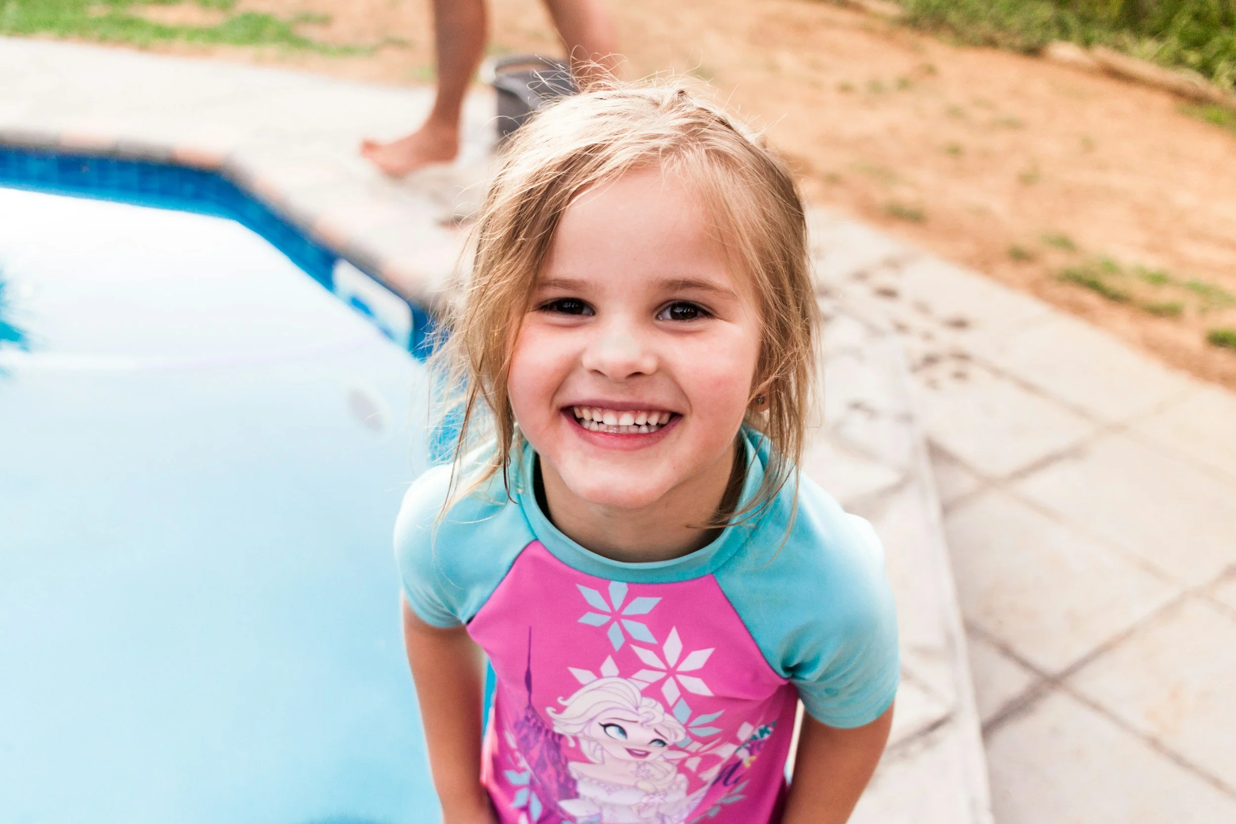 A young girl with blonde hair and a big smile, wearing a colorful swimsuit with a cartoon character, standing next to a swimming pool.