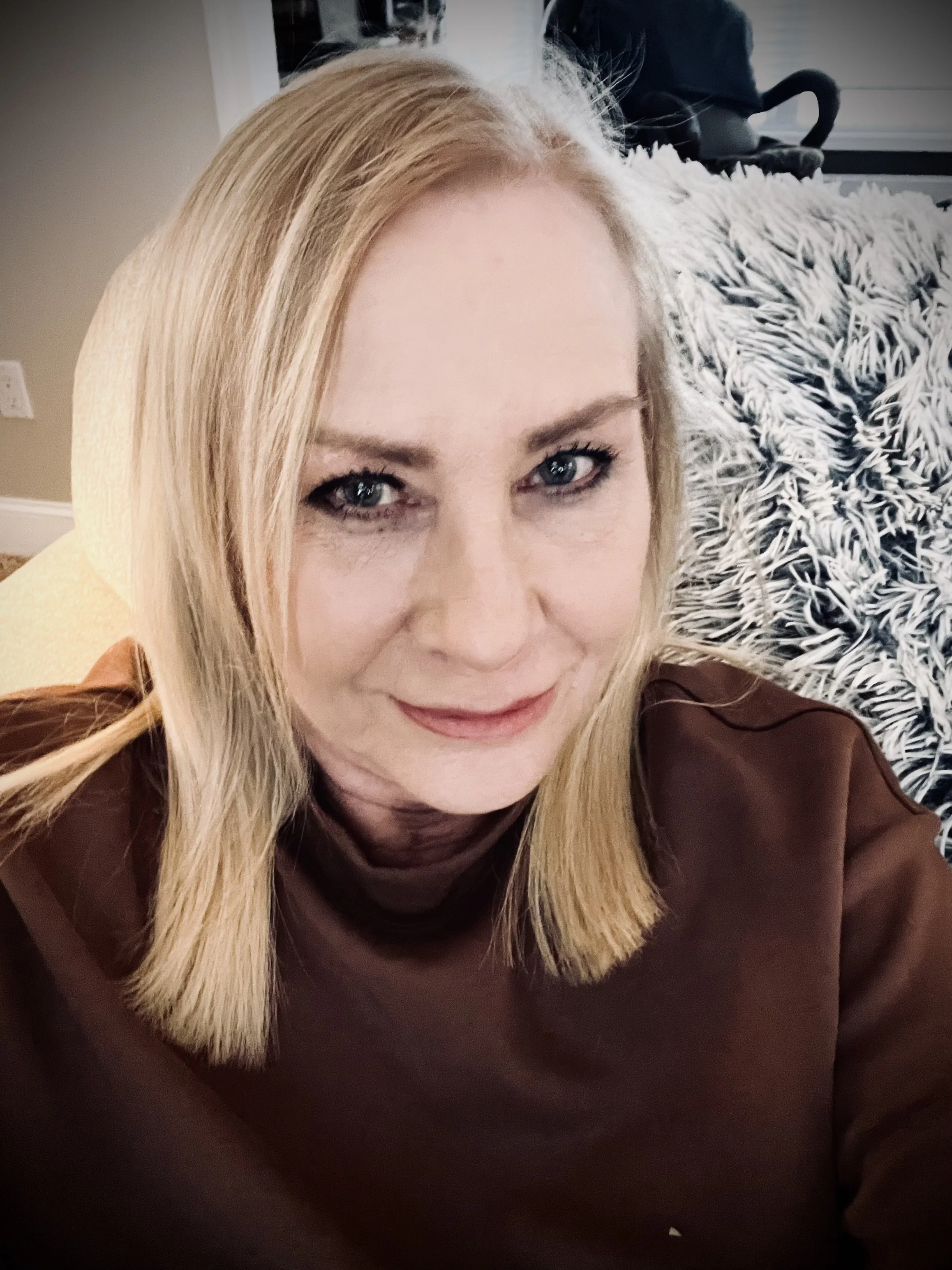 A woman with blonde hair and blue eyes sitting on a beige chair, smiling at the camera, with a black and white patterned blanket and a black backpack in the background.