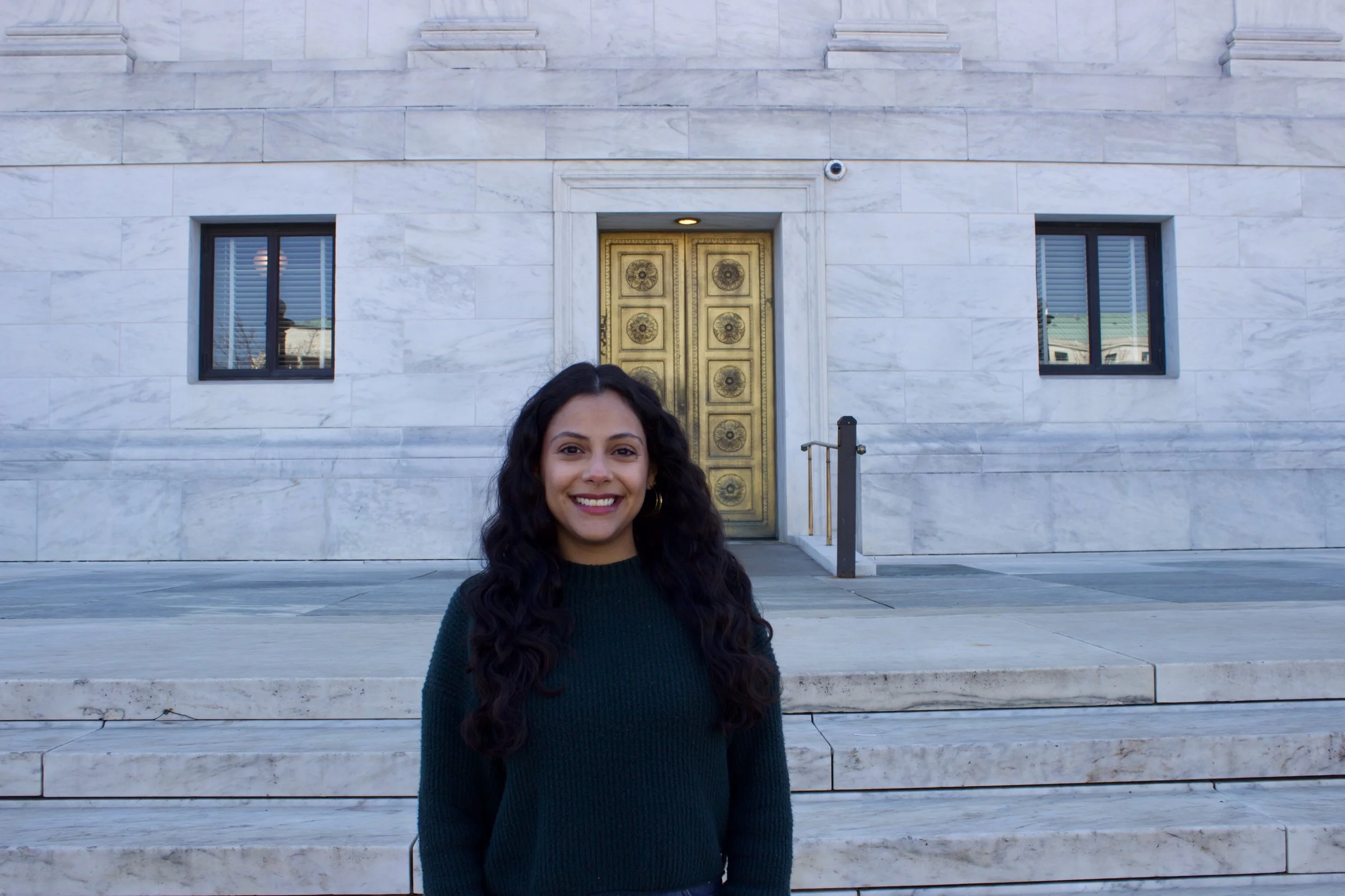 Young woman with dark curly hair smiling outside in front of a marble building with gold double doors and two small windows.