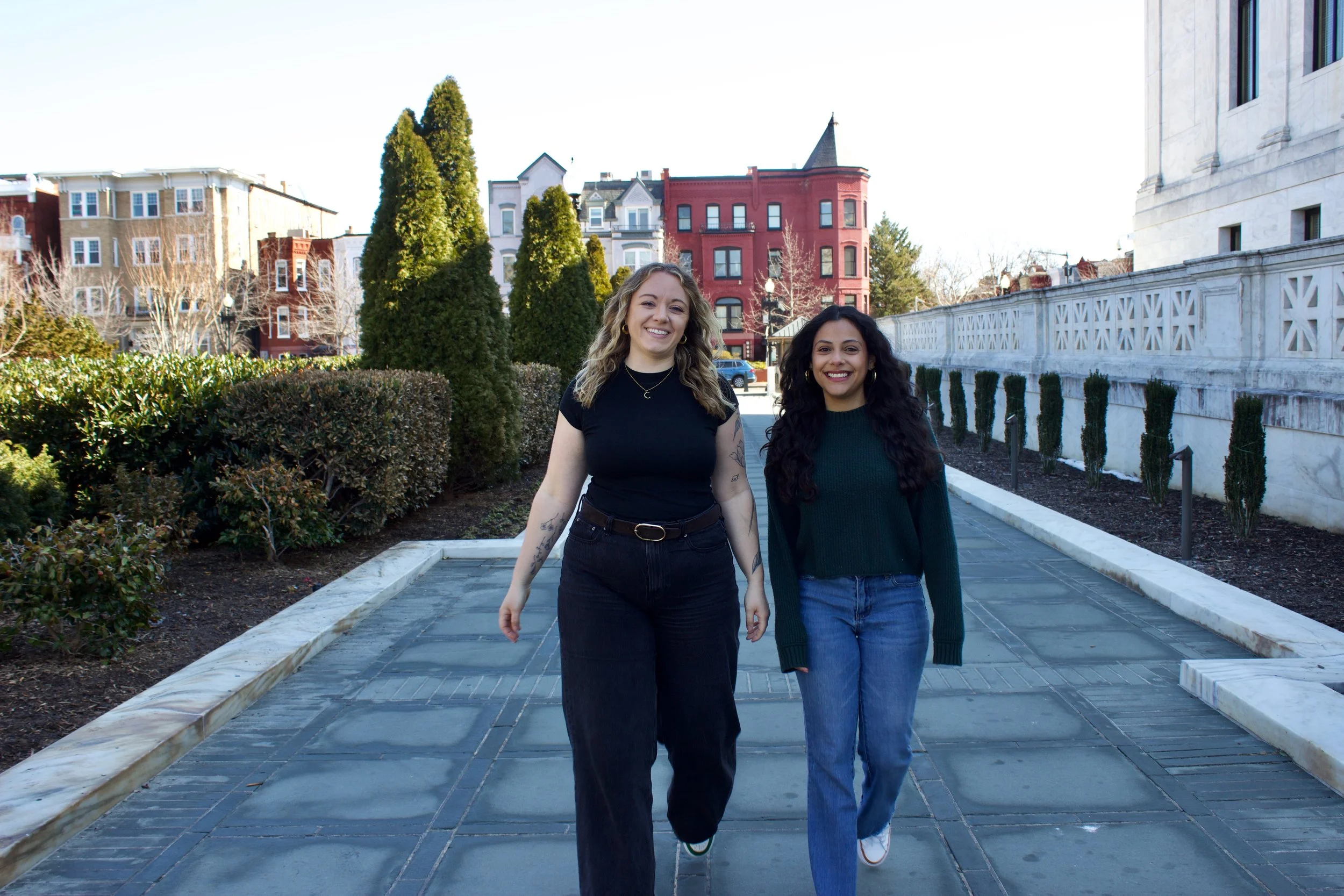 Two women walking along a paved path in an urban neighborhood with colorful Victorian-style houses and green trees in the background.