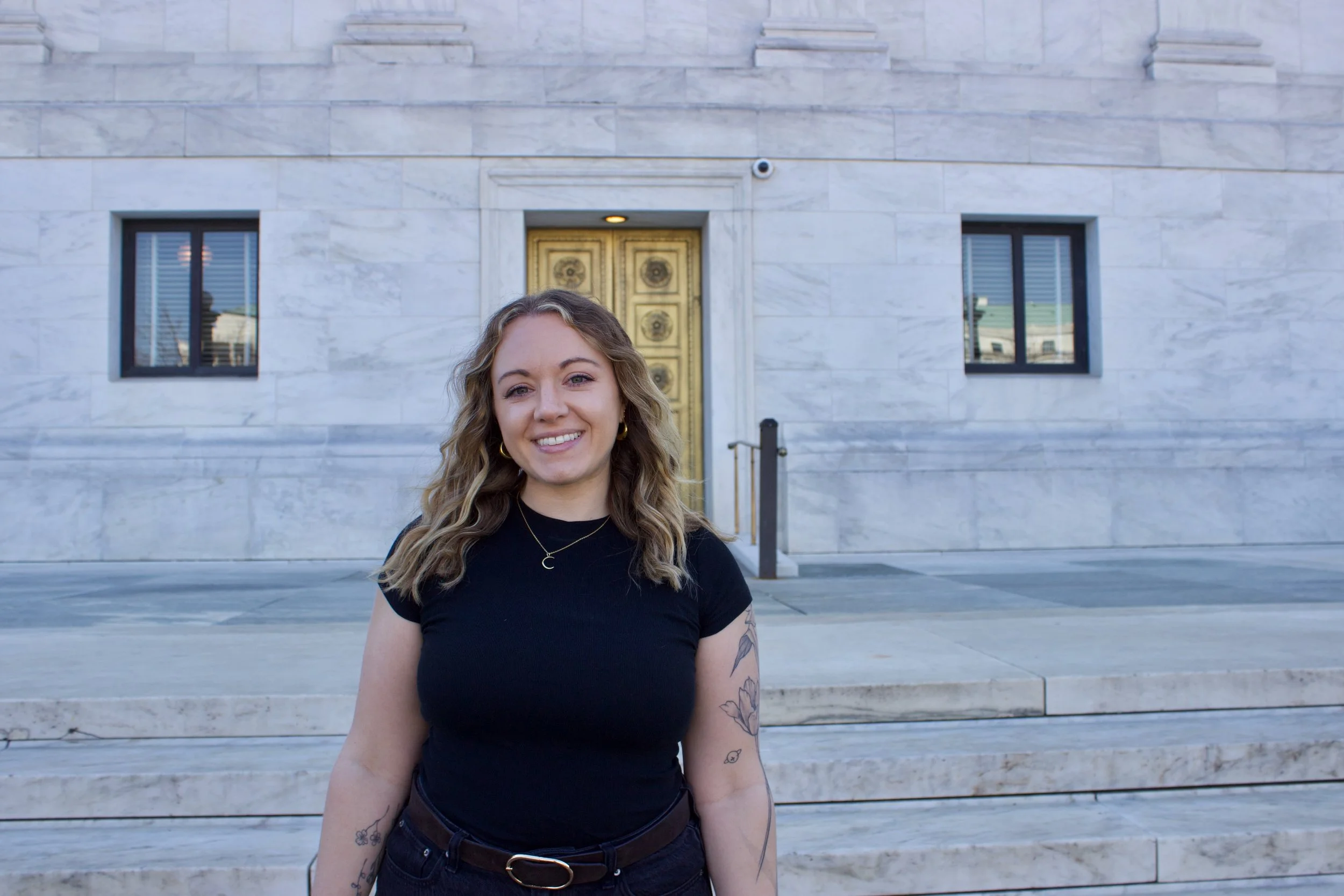 A woman with shoulder-length wavy hair, wearing a black t-shirt and jewelry, smiling outdoors in front of a white marble building with gold door and black-framed windows.