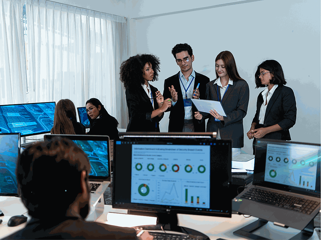 Group of five professionals in business attire having a discussion in a modern office with multiple computer screens displaying charts and data.