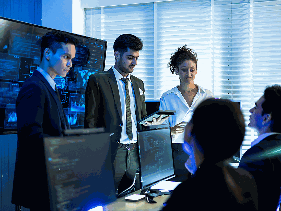 A diverse group of five professionals in an office, engaged in a serious discussion around a computer monitor, with digital data and graphs displayed on screens in the background.