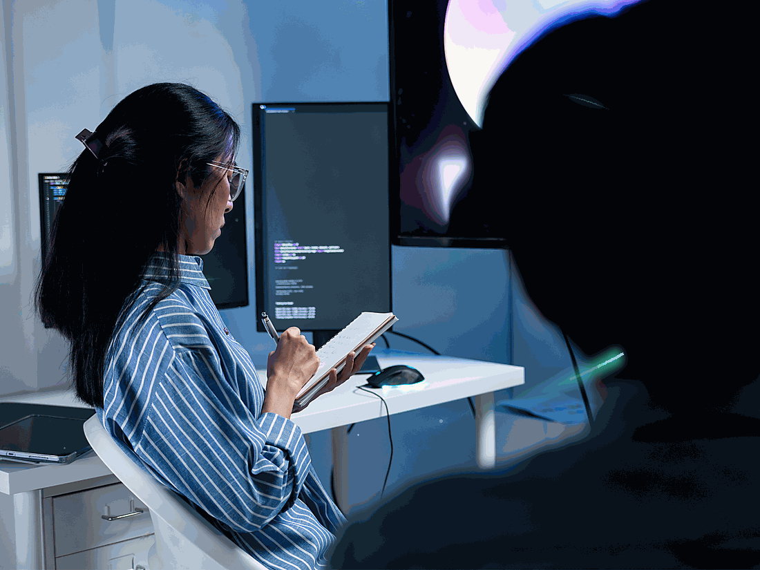 Woman with glasses in striped shirt seated at desk, writing in notebook, computer screens with code in the background in a modern office.