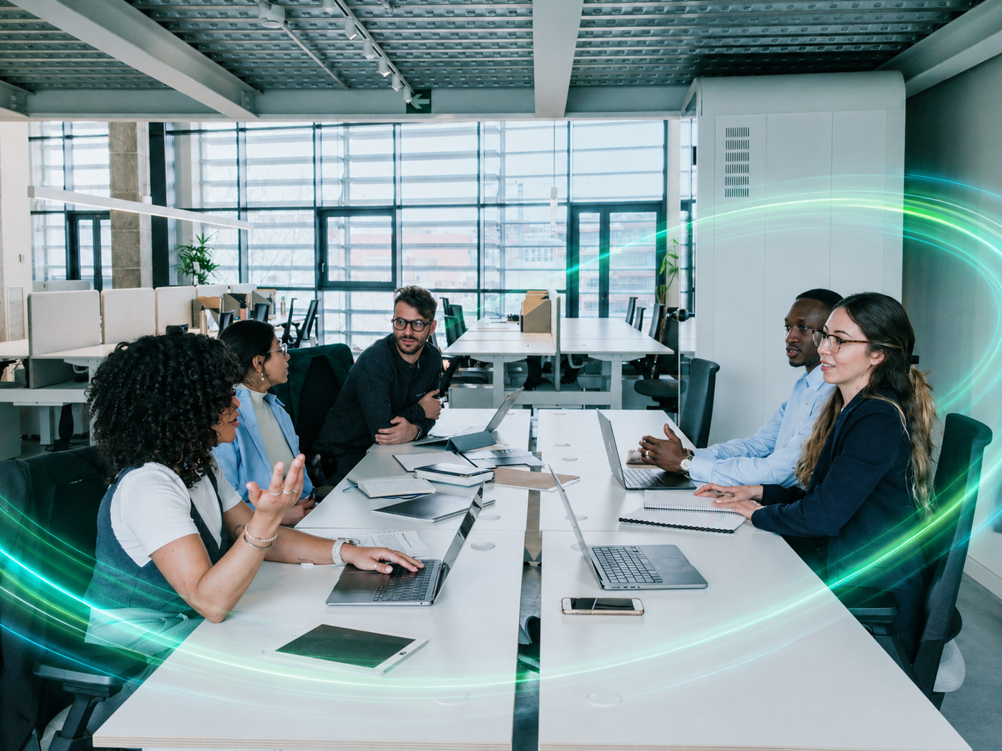 Six diverse professionals having a meeting around a table in a modern office, with laptops, tablets, and notebooks, engaging in discussion.
