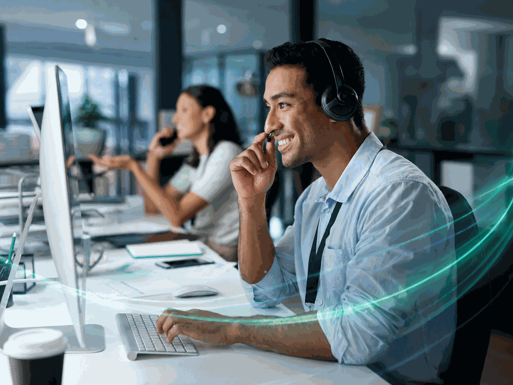 Man in an office wearing a headset working at a computer, smiling. Woman in background on the phone.