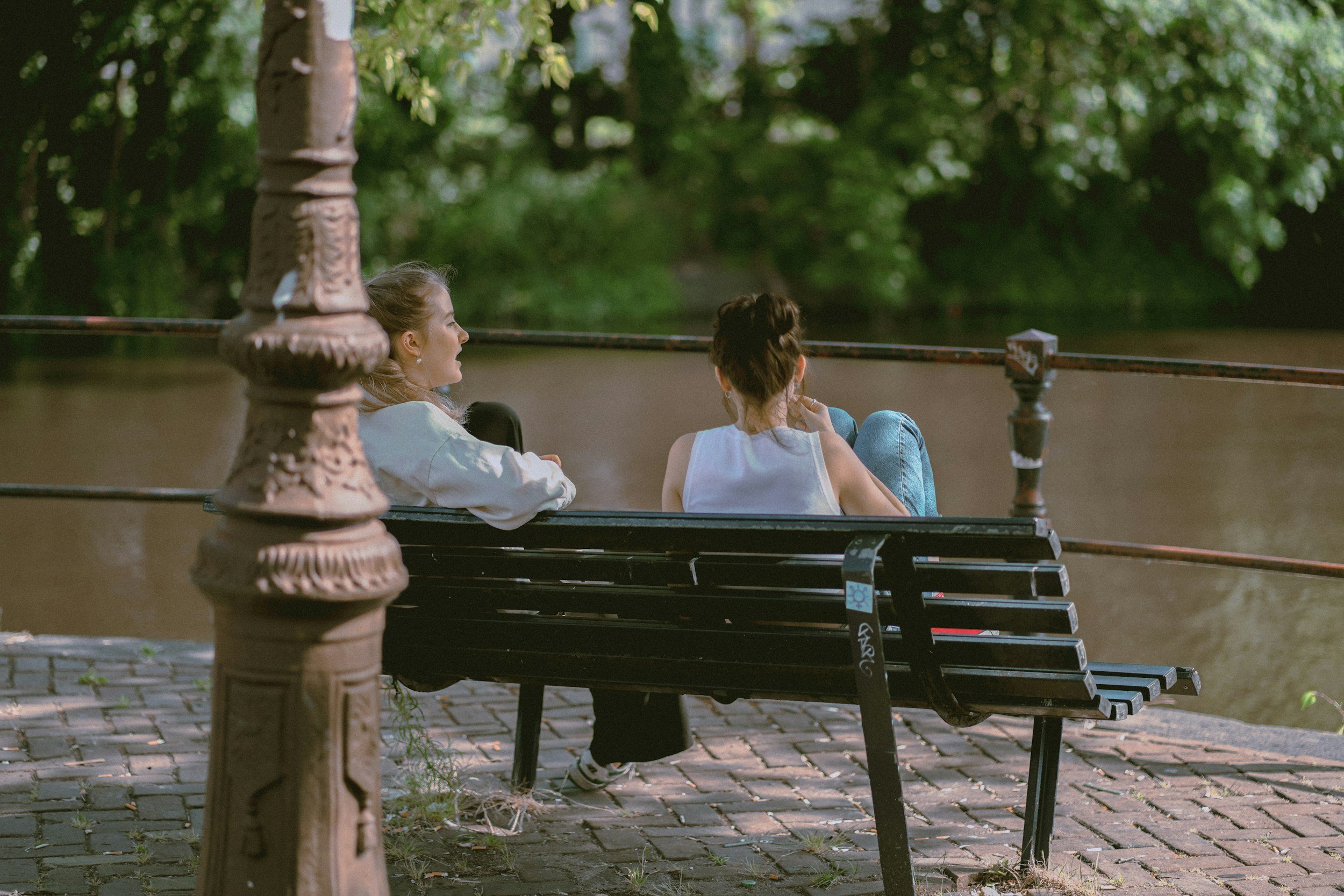 Two women sit on a park bench near a river, engaged in conversation. One woman faces the river, while the other looks at her. The park is lush with green trees.