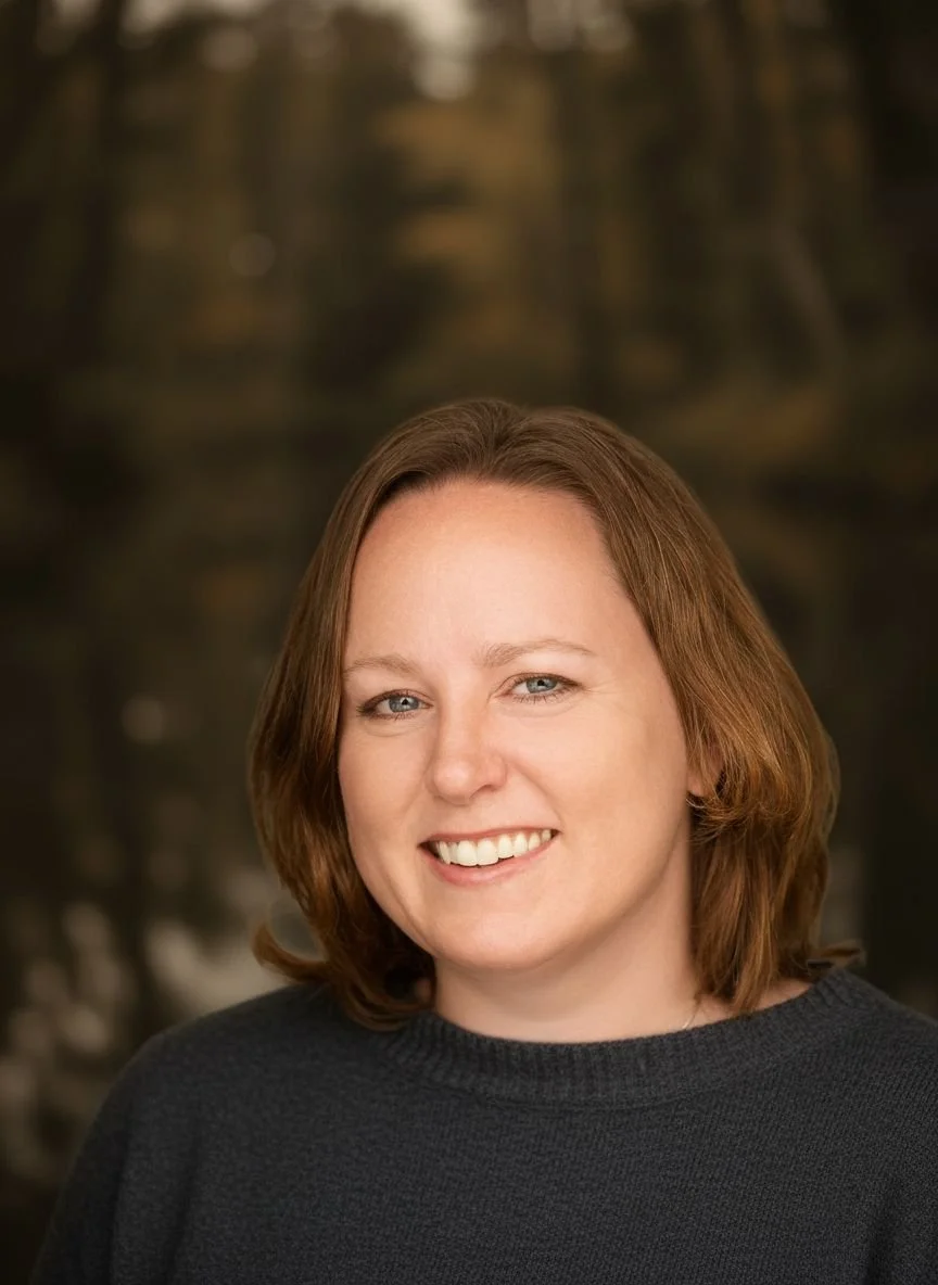 A woman with shoulder-length brown hair, blue eyes, and a bright smile, wearing a dark sweater, outdoors in a blurred natural setting.
