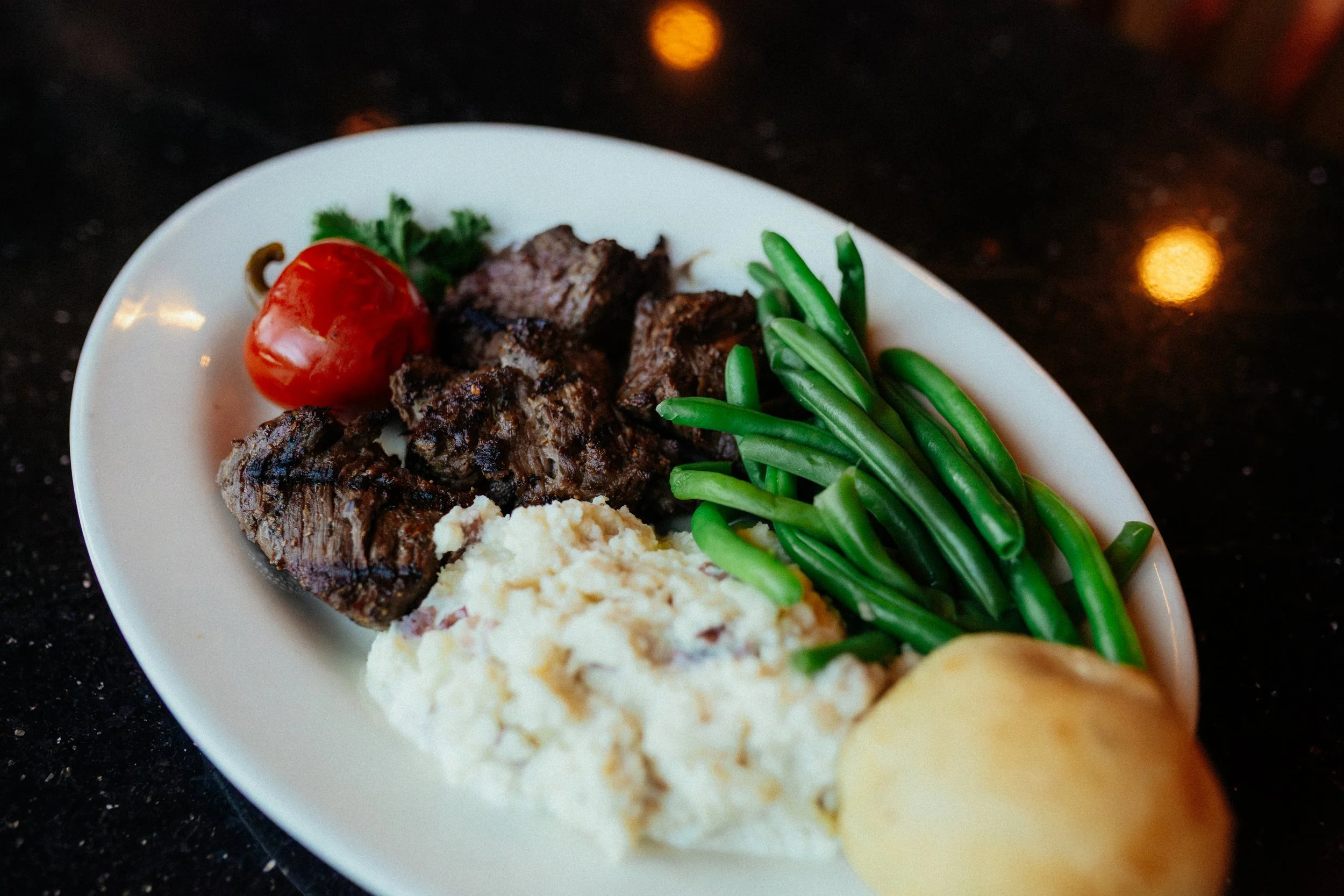 Plate of grilled beef chunks, a cherry tomato, green beans, mashed potatoes with gravy, and a bread roll on a black countertop