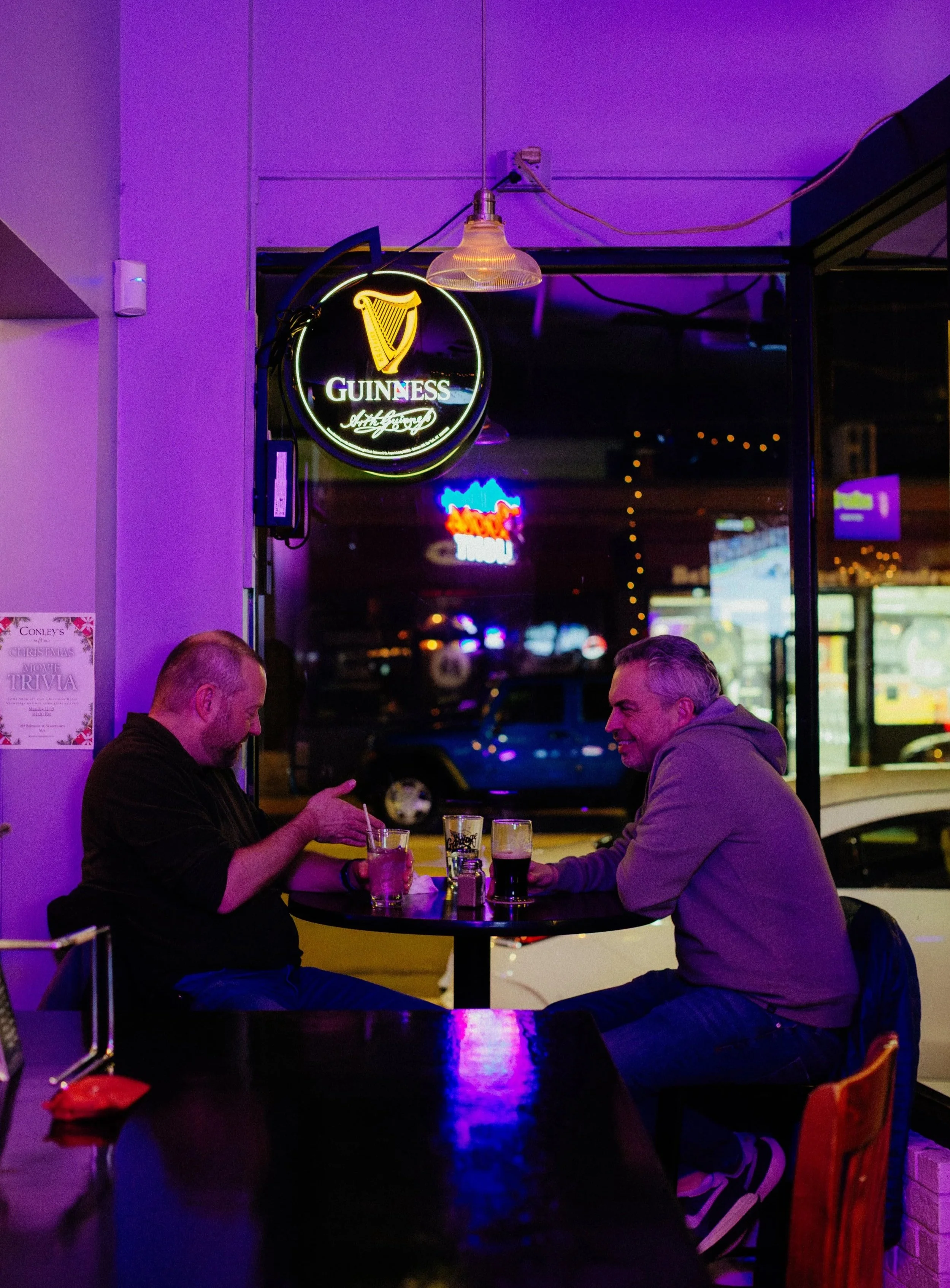 Two men sitting at a table in a bar, enjoying drinks, with neon signs and a window showing a nighttime street scene in the background.