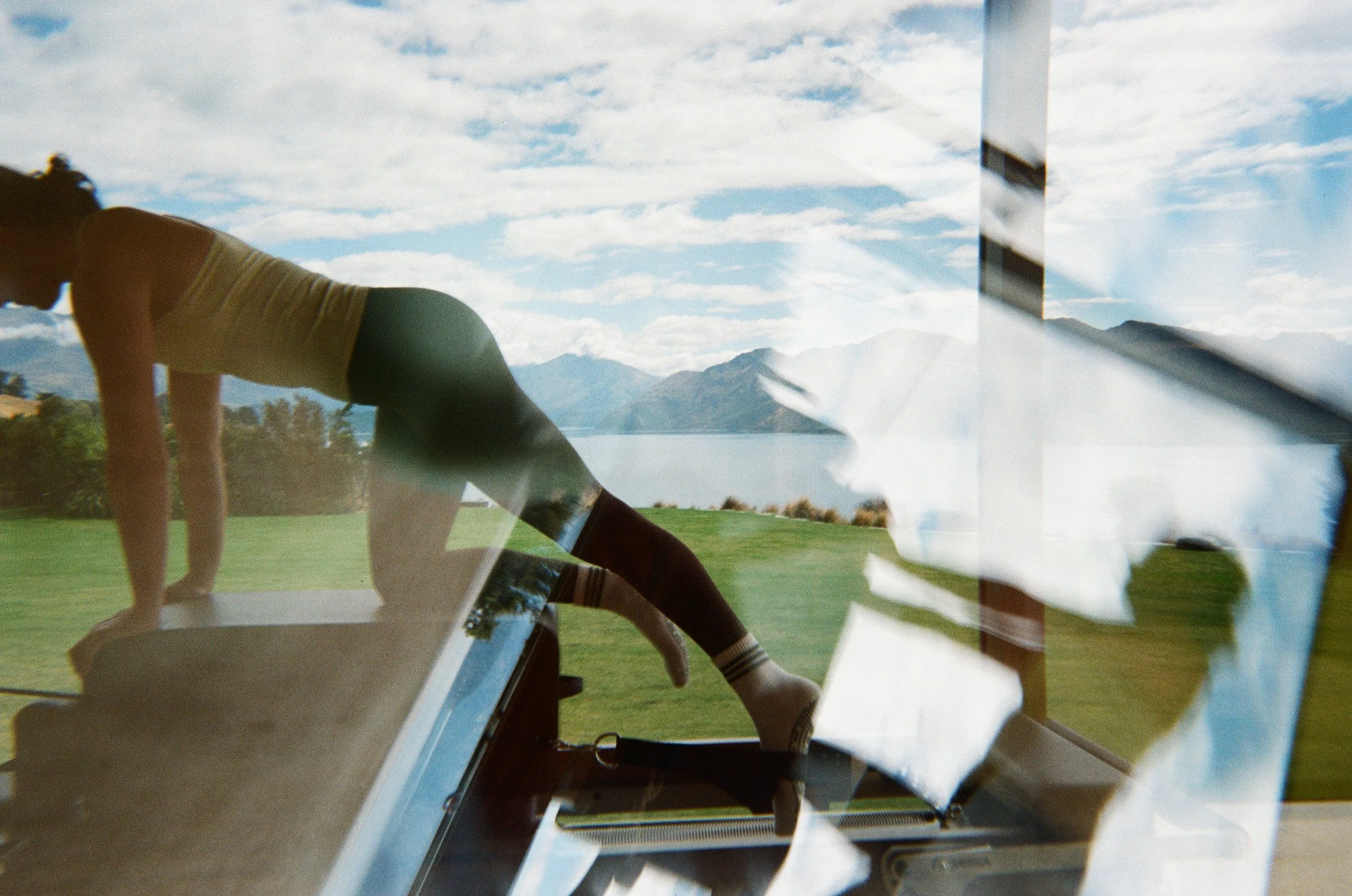 Reflection of a person doing yoga outdoors, with mountains, lake, cloudy sky, and a window with paper notes reflected in the glass.