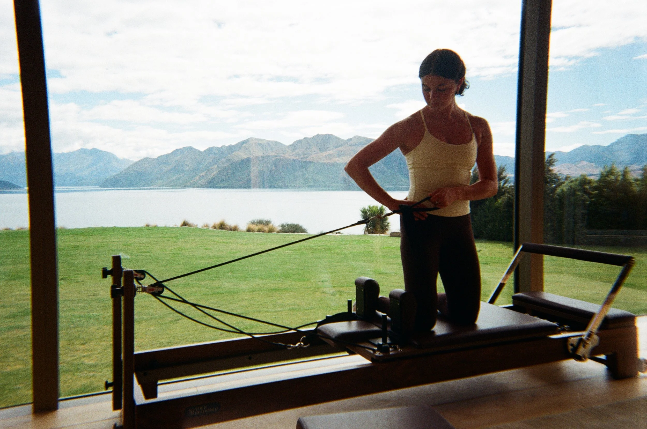 A woman using a Pilates reformer machine indoors with a large window showing mountains and a lake outside.