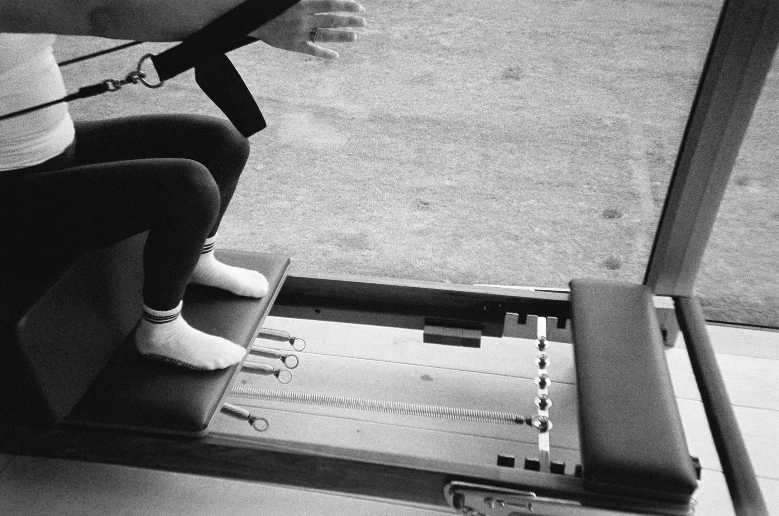 A person wearing white socks and black leggings is seated on a Pilates reformer machine, holding onto a strap with one hand, in a studio with a textured floor.