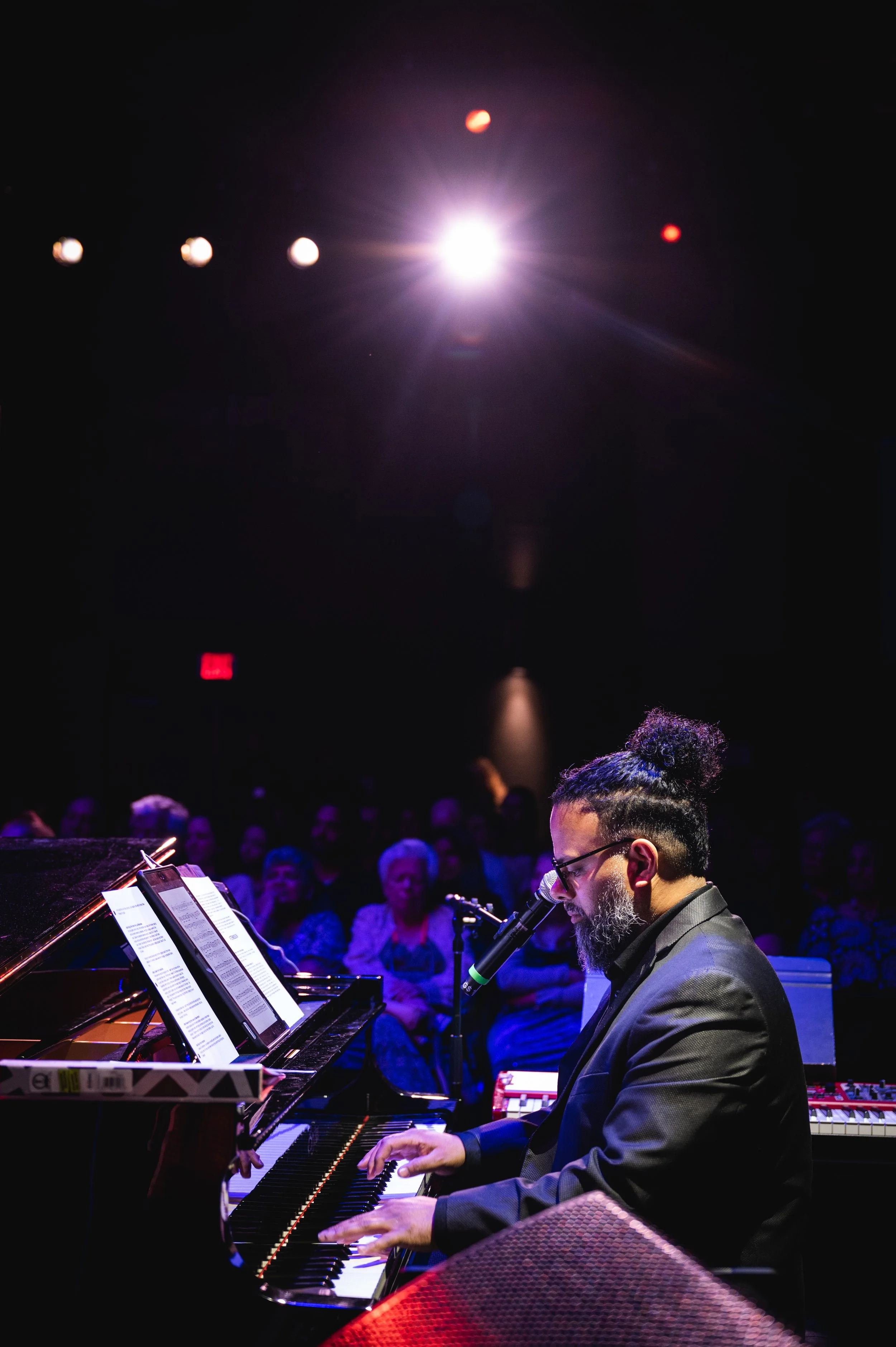 Musician performing live at a grand piano under stage lighting before an audience.