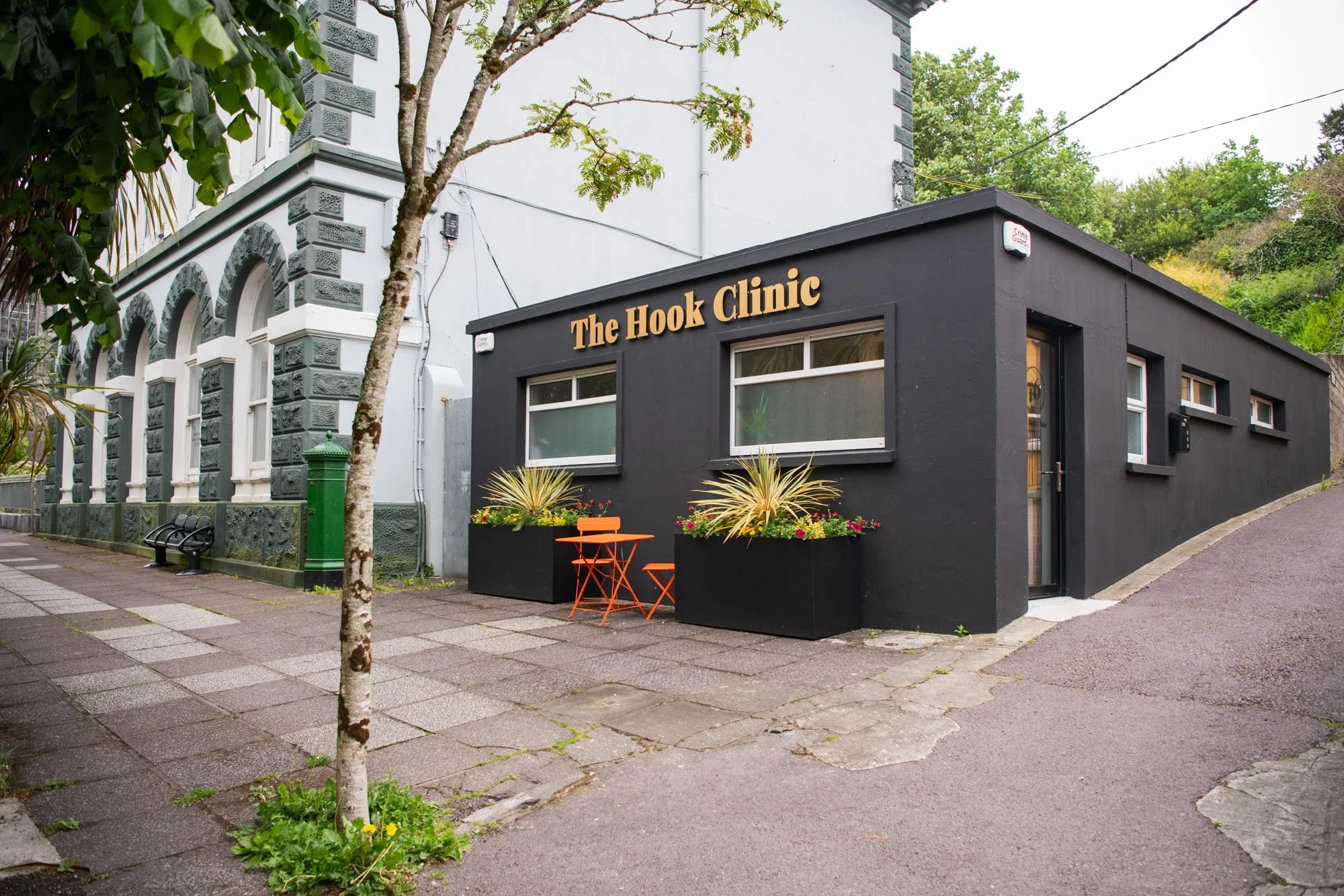 Exterior of a small black building labeled 'The Hook Clinic' with orange outdoor furniture and plants outside, situated on a sidewalk next to a larger white historic-style building with arched windows, surrounded by trees and greenery.