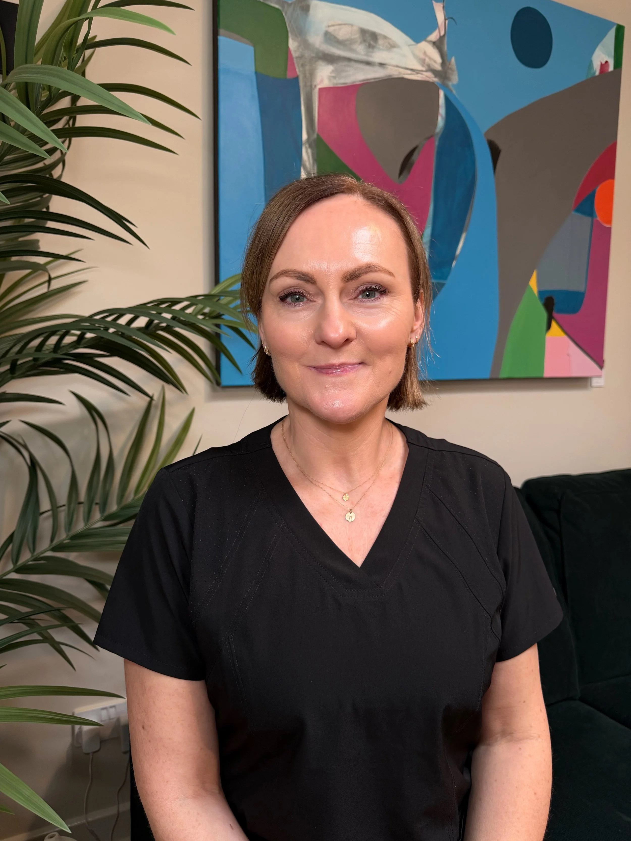 A woman with short brown hair and blue eyes smiling slightly, wearing a black top and gold jewelry, sitting in front of an abstract colorful painting and a large green plant.