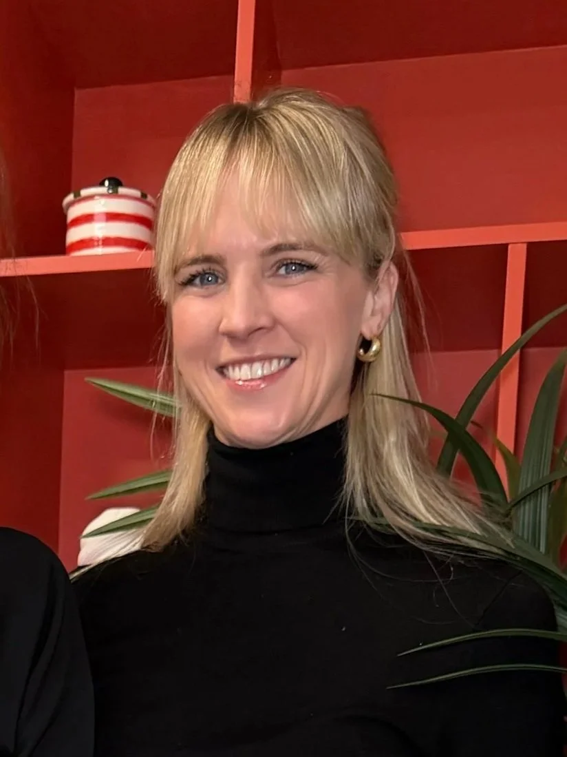 A smiling woman with blonde hair, wearing a black turtleneck and gold earrings, standing in front of a red shelf with a plant and a red and white striped container.