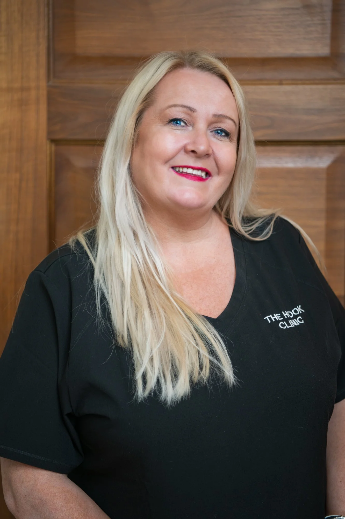 A woman with long blonde hair and blue eyes smiling, wearing a black scrub top with the text 'The Hook Clinic' on it, standing against a wooden background.