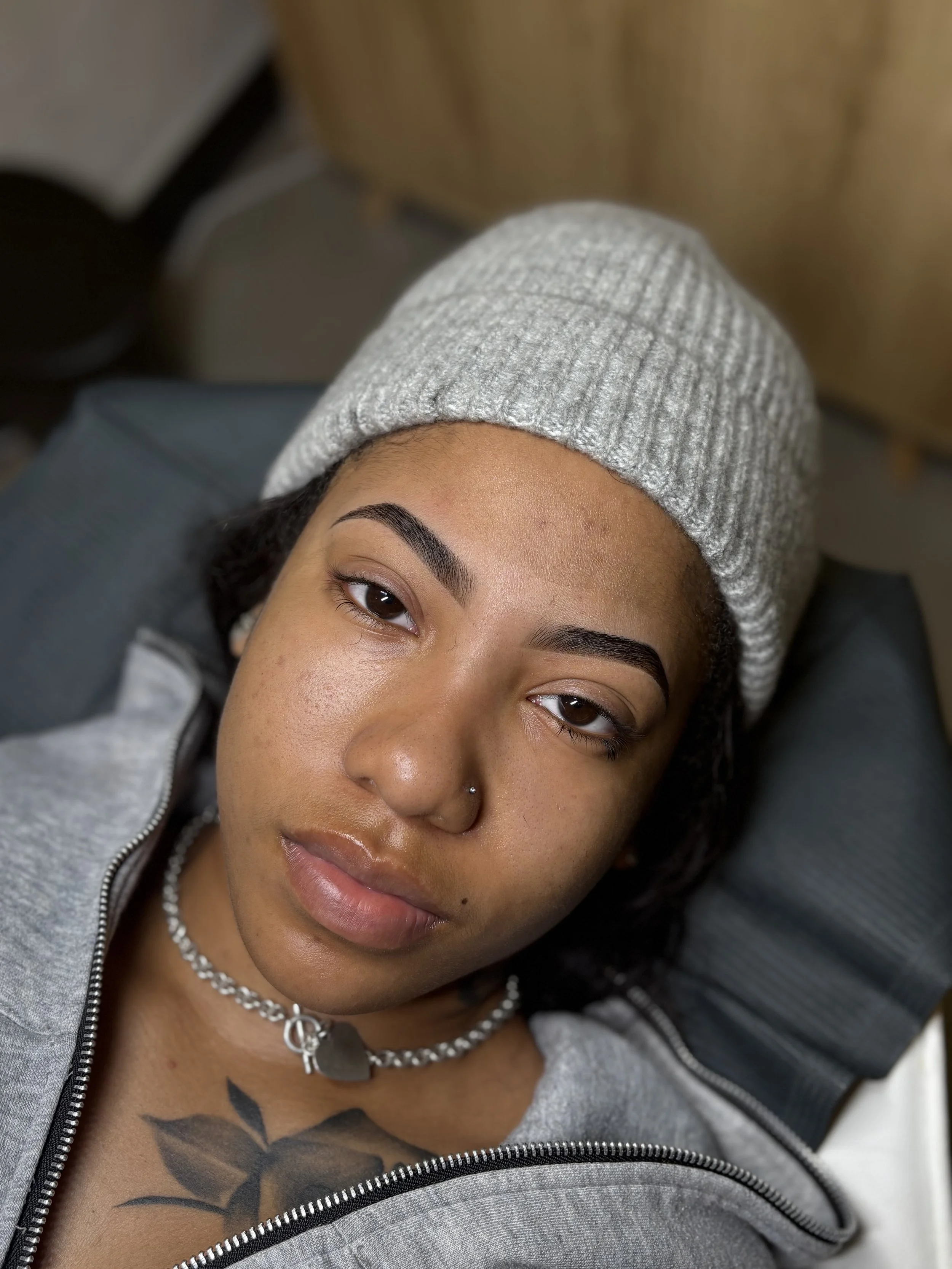 A young woman with a nose piercing, wearing a gray knit beanie, lying down with a relaxed expression, wearing a gray zip-up jacket and necklace, showing a tattoo on her chest.